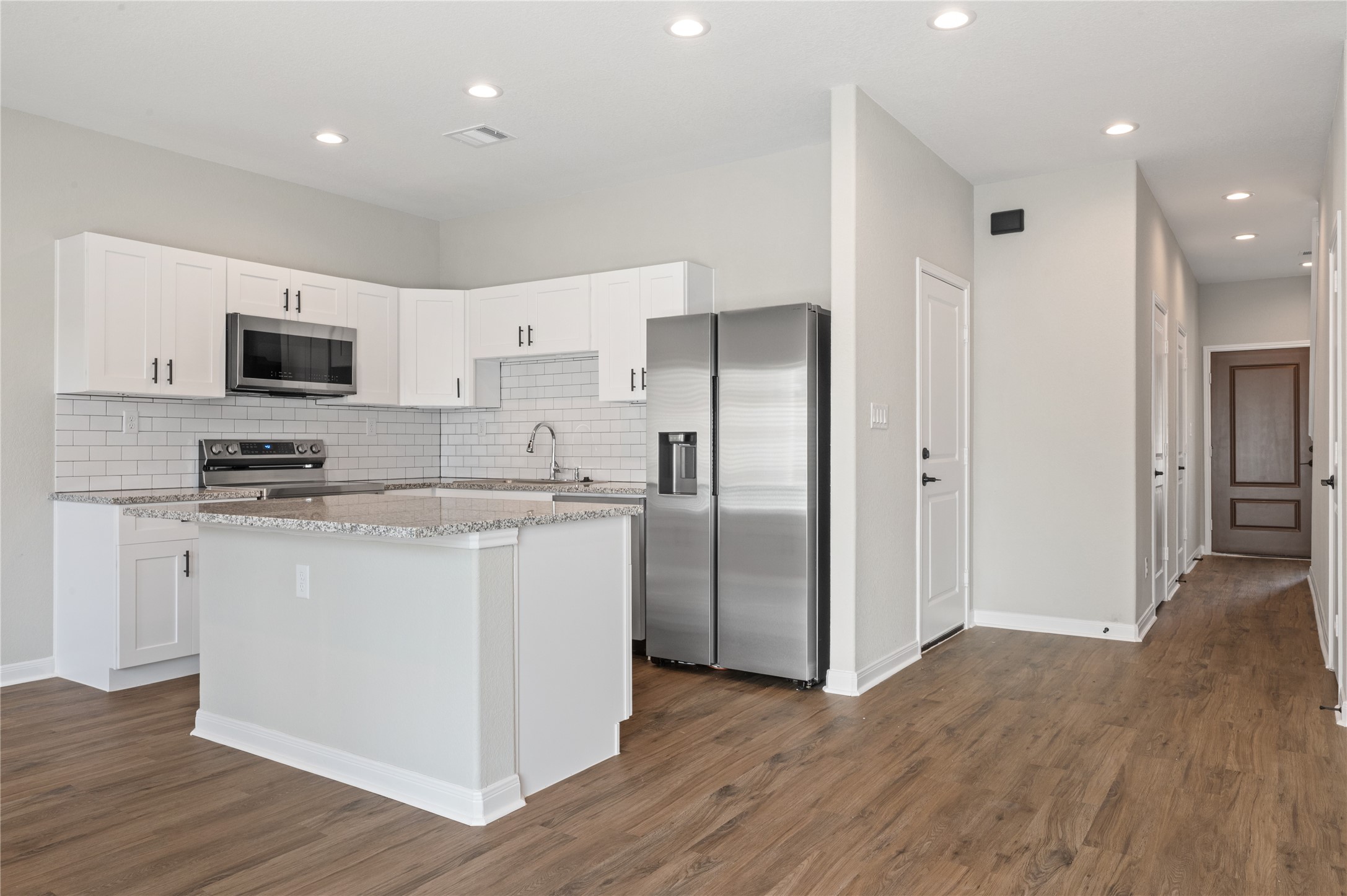 14010 Longstreet Road, Unit 2 Willis, TX 77318 - Photo 23 of 29 This modern kitchen features white cabinetry, stainless steel appliances, and granite countertops. It includes a central island and a subway tile backsplash. The open layout with wood-look flooring leads into a hallway, creating a spacious and inviting atmosphere.