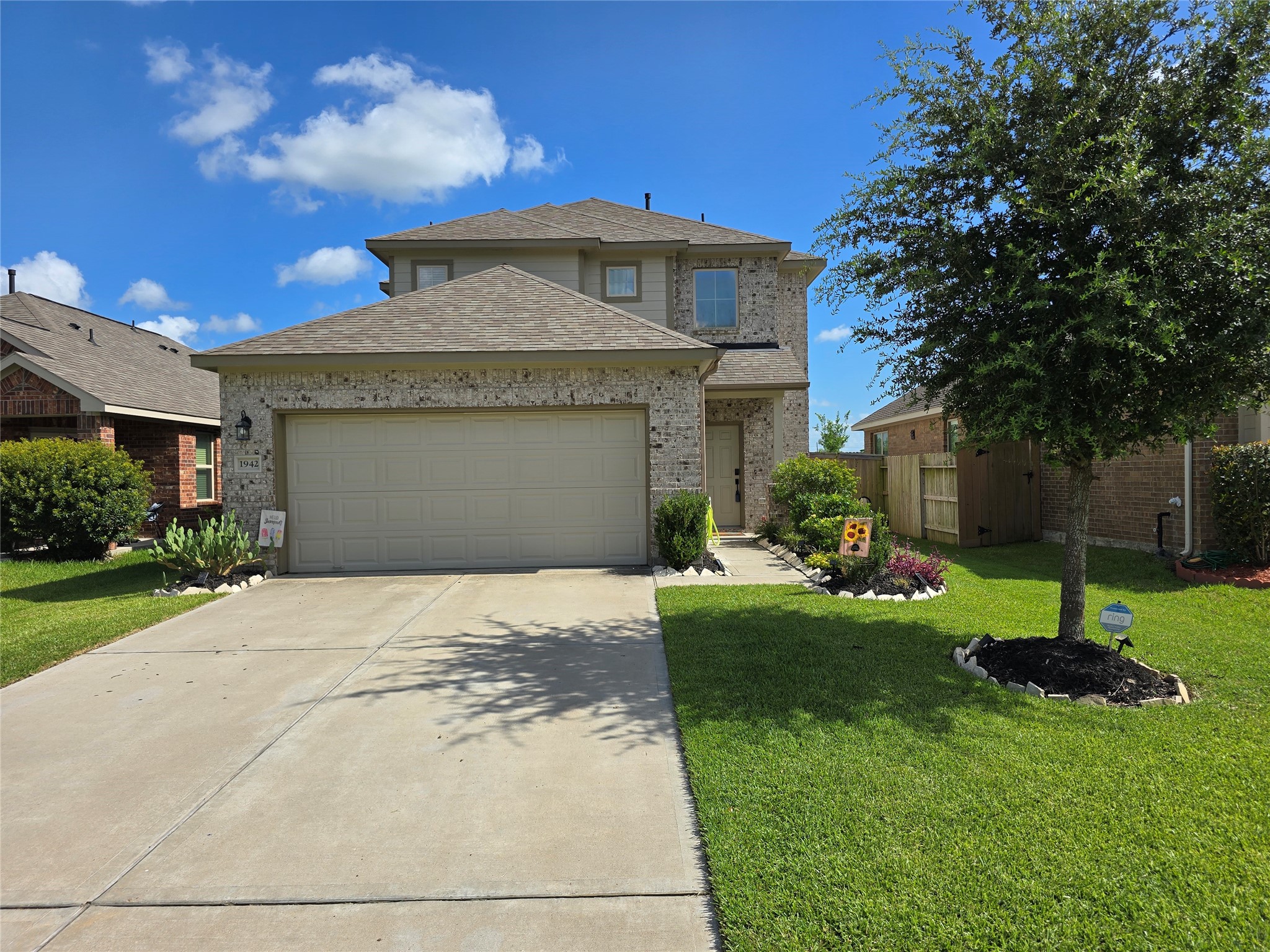 This photo showcases a two-story brick home with a two-car garage. The property features a well-maintained front lawn, landscaped garden beds, and a mature tree. It is set in a suburban neighborhood with similar homes nearby.