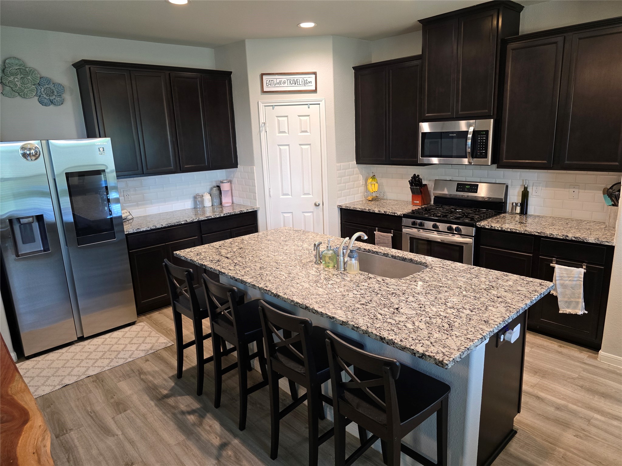 1942 Bending Green Drive Rosharon, TX 77583 - Photo 11 of 30 This modern kitchen features dark wood cabinets, granite countertops, and stainless steel appliances. The spacious island includes a sink and seating area, perfect for casual dining. The room is well-lit with recessed lighting and has a sleek, contemporary design.