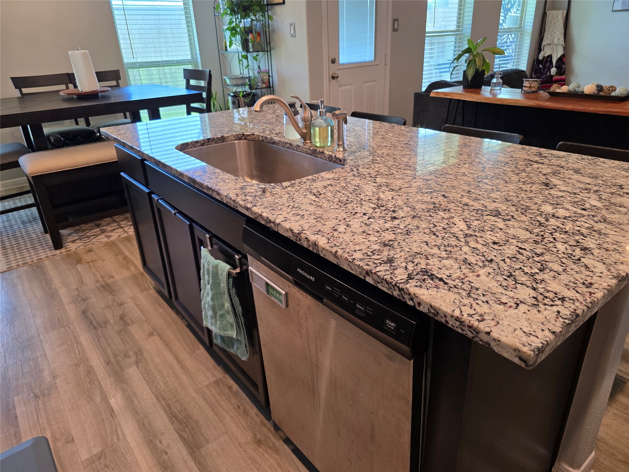 1942 Bending Green Drive Rosharon, TX 77583 - Photo 13 of 30 This kitchen features a spacious granite island with a sink and dishwasher, complemented by dark cabinetry. The open layout includes a dining area with natural light, hardwood floors, and modern decor, creating a warm and inviting atmosphere.