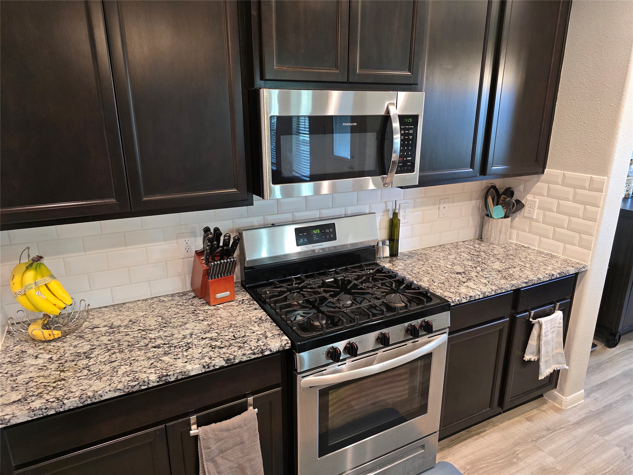 1942 Bending Green Drive Rosharon, TX 77583 - Photo 14 of 30 Modern kitchen with dark wood cabinets, granite countertops, and stainless steel appliances, including a gas stove and microwave. Subway tile backsplash and light wood flooring add a stylish touch.