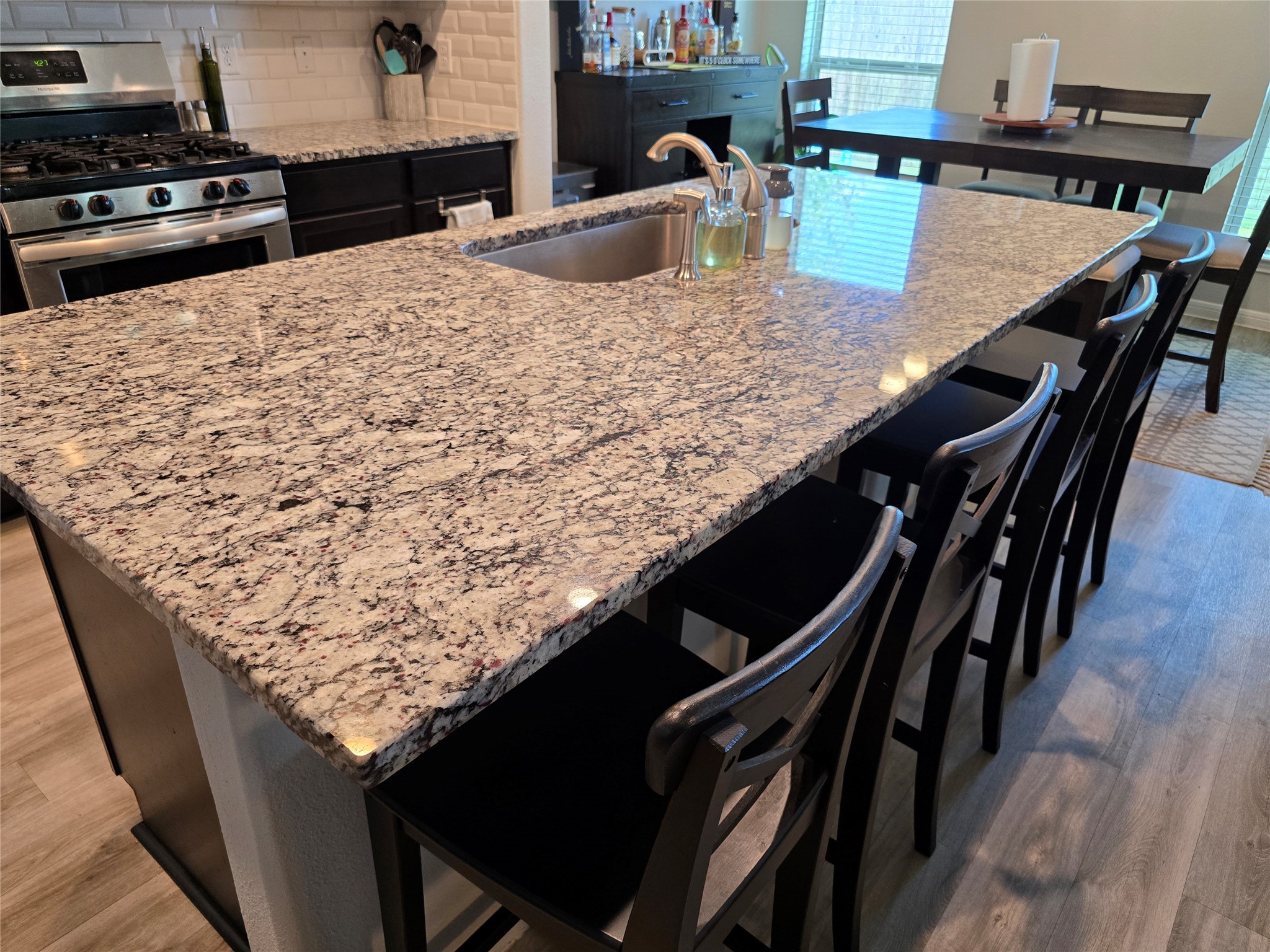 1942 Bending Green Drive Rosharon, TX 77583 - Photo 15 of 30 This modern kitchen features a large granite island with a built-in sink, perfect for meal preparation and casual dining. The space includes a stainless steel gas stove, dark cabinetry, and a dining area that accommodates several people, enhanced by natural light from nearby windows.
