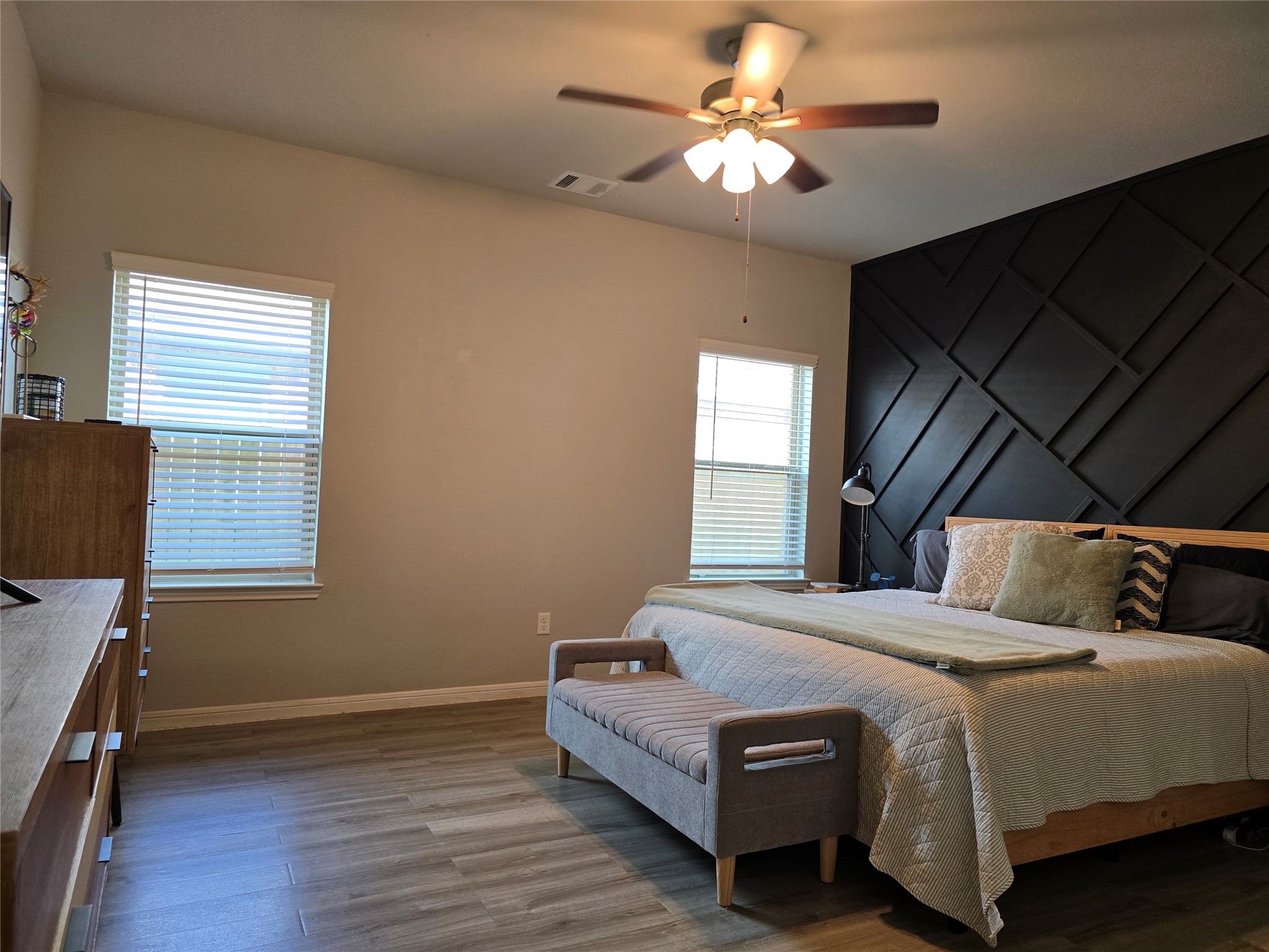 1942 Bending Green Drive Rosharon, TX 77583 - Photo 16 of 30 This photo showcases a spacious bedroom with a modern design. It features a large bed, a textured black accent wall, wood flooring, and a ceiling fan. Two windows provide ample natural light, creating a bright and inviting atmosphere.