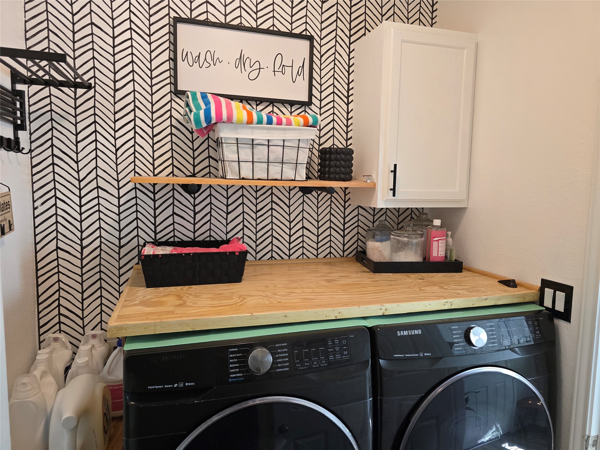 1942 Bending Green Drive Rosharon, TX 77583 - Photo 27 of 30 This photo shows a stylish laundry room with a modern black and white herringbone accent wall. It features a washer and dryer set under a wooden countertop, with shelves for storage, a cabinet, and decorative elements for an organized and functional space.
