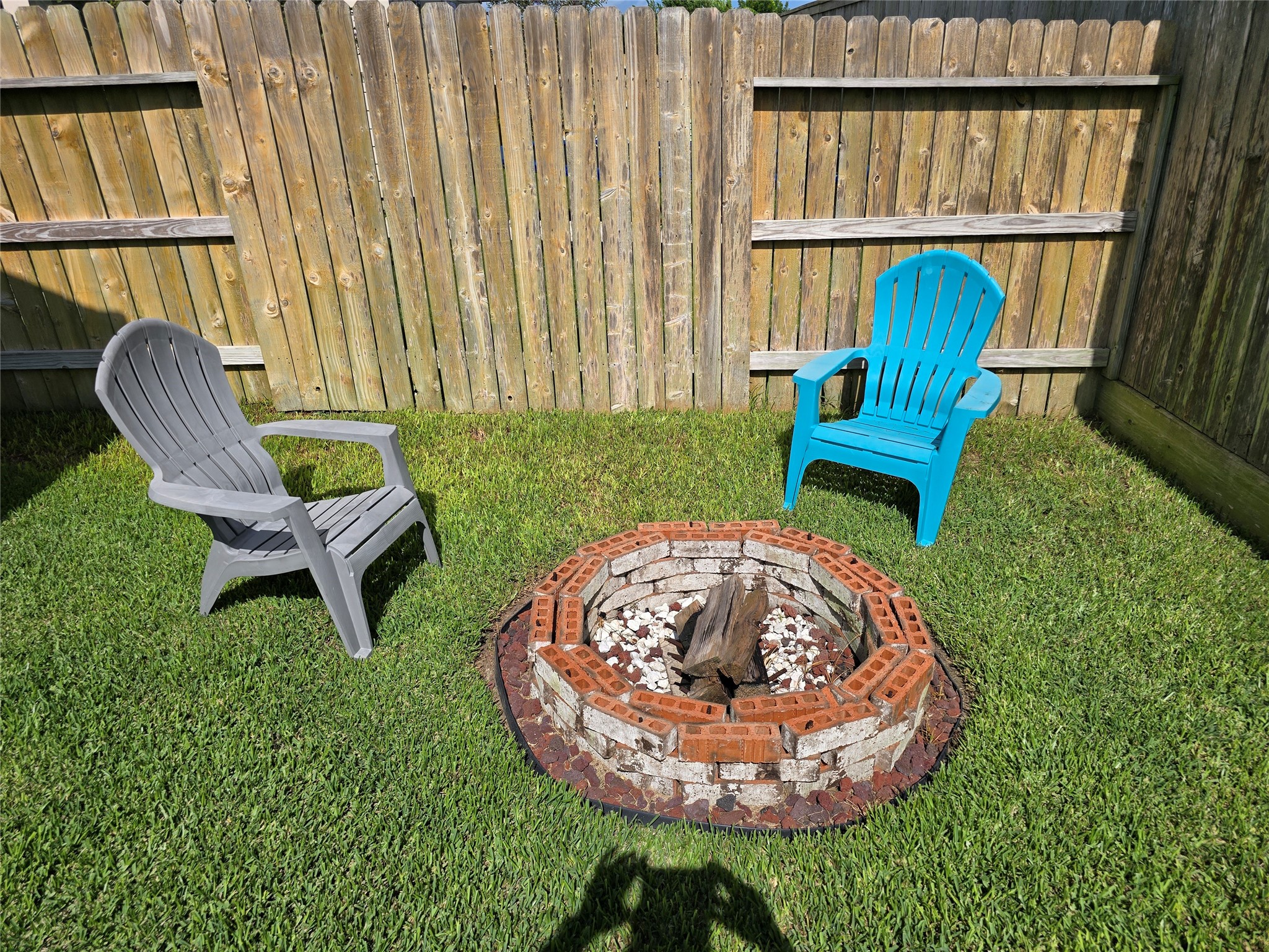 1942 Bending Green Drive Rosharon, TX 77583 - Photo 28 of 30 This photo shows a cozy backyard corner featuring a fire pit surrounded by a wooden fence. Two colorful Adirondack chairs are placed on the well-maintained grass, creating a perfect spot for outdoor relaxation.