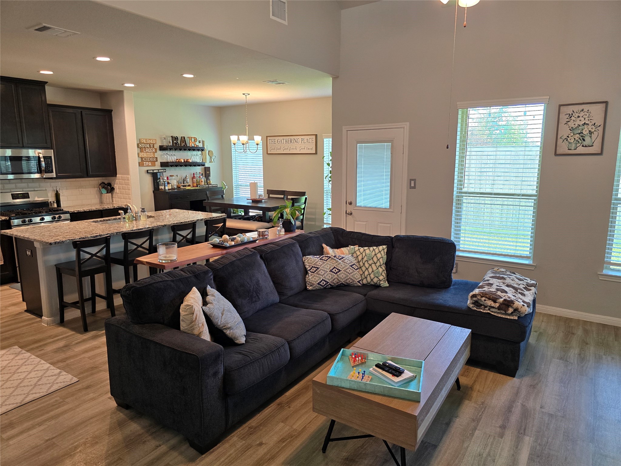 1942 Bending Green Drive Rosharon, TX 77583 - Photo 4 of 30 This inviting open-concept living space features a modern kitchen with dark cabinetry, a spacious island with bar seating, and a cozy living area with a large sectional sofa. Natural light fills the room through multiple windows, creating a bright and welcoming atmosphere.