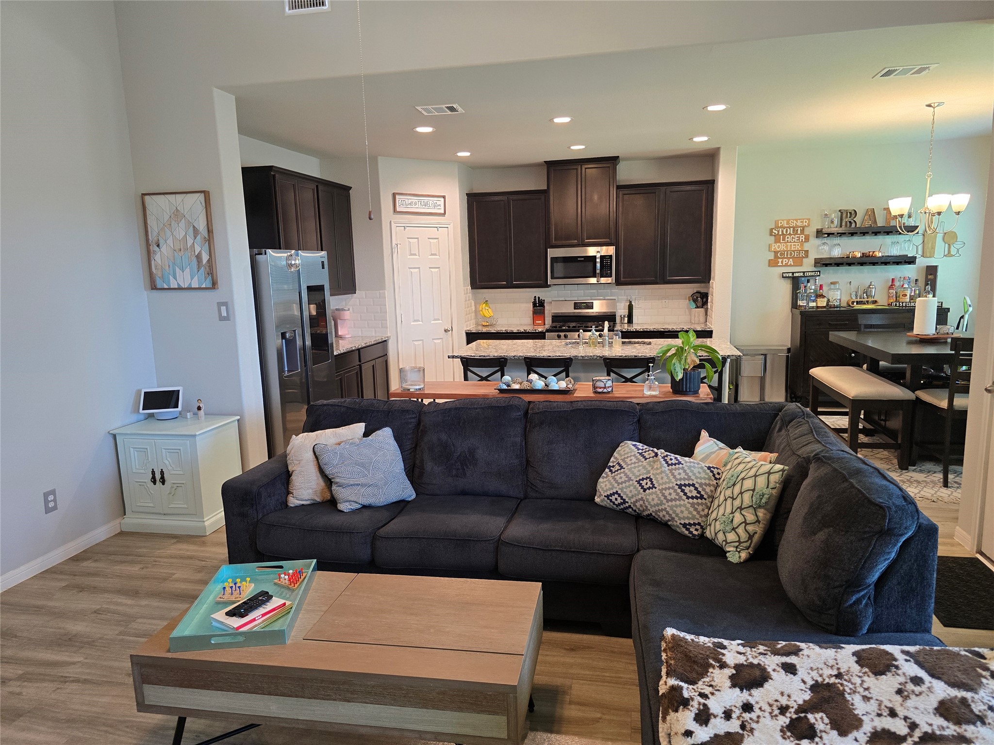 1942 Bending Green Drive Rosharon, TX 77583 - Photo 5 of 30 This inviting open-concept space features a modern kitchen with dark cabinets and stainless steel appliances, seamlessly flowing into a cozy living area with a plush sectional sofa. A dining area with a stylish light fixture completes the setting, offering a perfect blend of comfort and functionality.