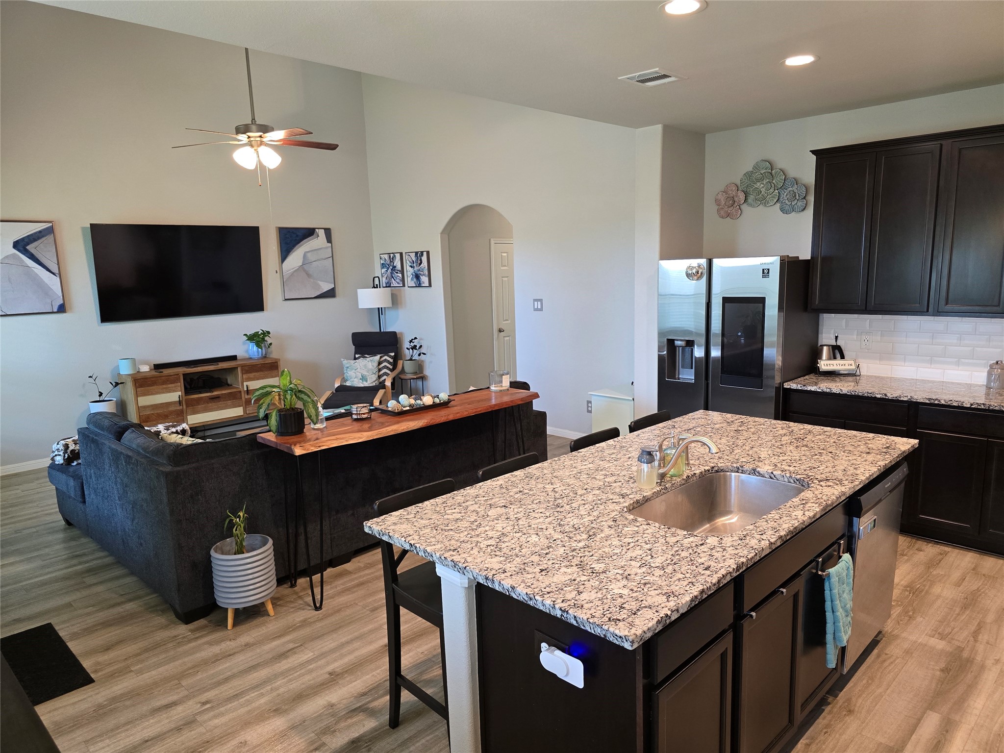 1942 Bending Green Drive Rosharon, TX 77583 - Photo 6 of 30 This photo showcases a modern open-concept living area with a spacious kitchen island featuring granite countertops and a sink. The room includes a cozy seating area, a wall-mounted TV, and stylish decor. The space is bright with a neutral color palette and hardwood flooring, offering a welcoming atmosphere perfect for entertaining.