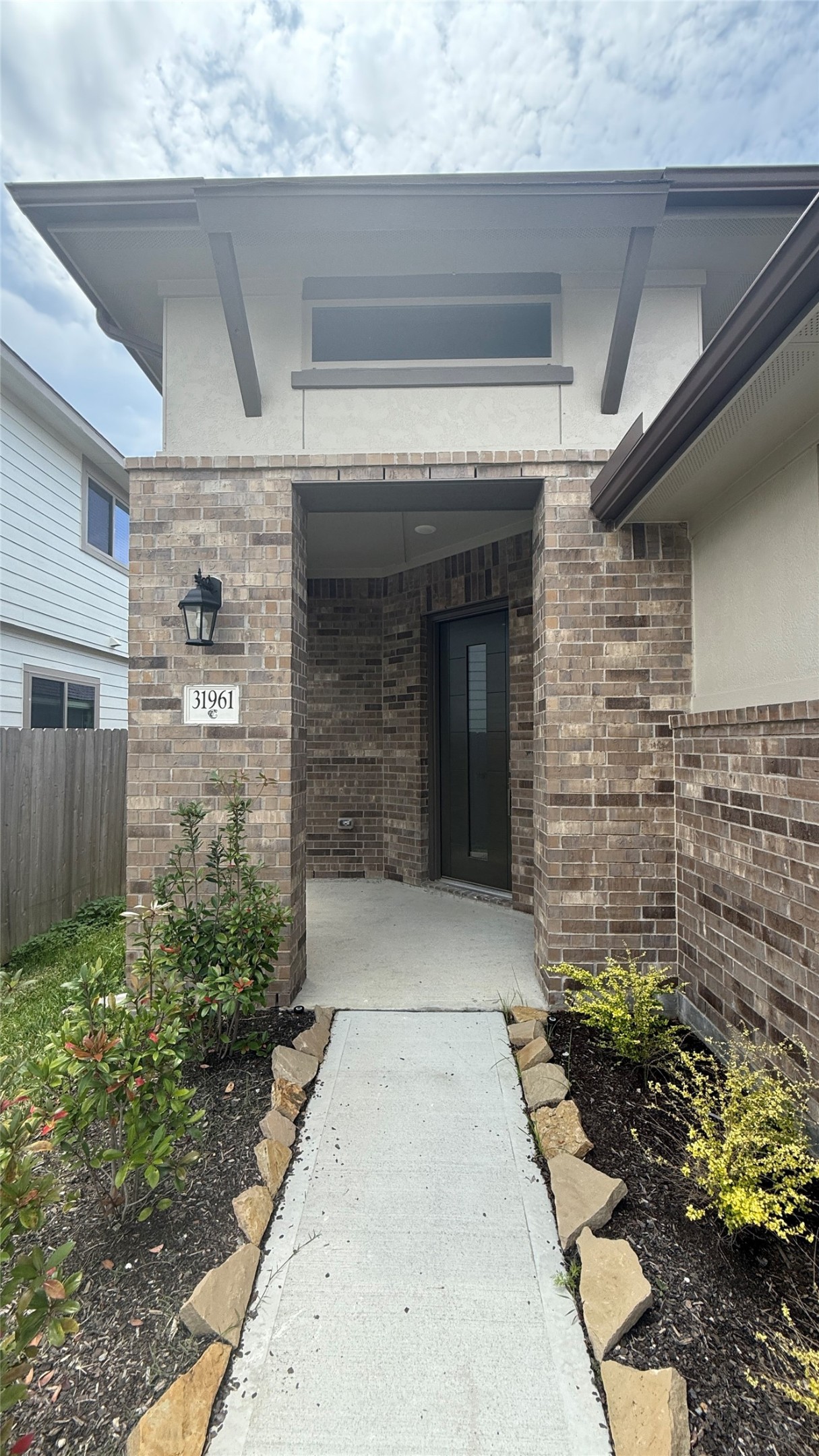 31961 Retama Rnch Drive Spring, TX 77386 - Photo 4 of 25 This photo showcases the entrance of a modern home with a brick exterior. A paved walkway with stone edging leads to a recessed front door framed by a covered porch. The surrounding landscaping includes low shrubs and plants, providing a welcoming and well-maintained appearance.