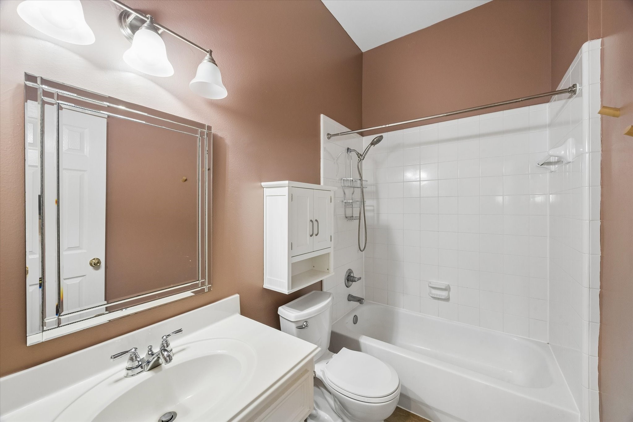 3118 Valley Court Manvel, TX 77578 - Photo 18 of 38 This is a clean, well-lit bathroom with white fixtures, including a bathtub with a shower, a toilet, and a sink. It features a large mirror, ample lighting, and a wall-mounted cabinet for storage. The walls are painted a warm beige.