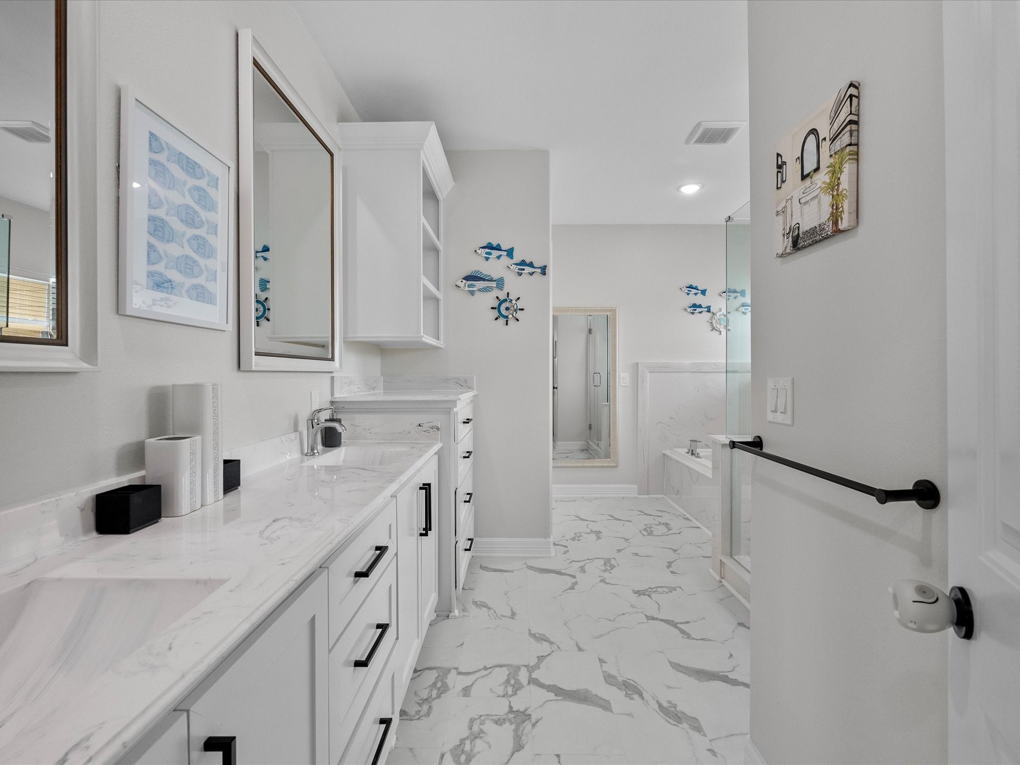 1018 Seashell Drive Surfside Beach, TX 77541 - Photo 43 of 49 This is a spacious, modern bathroom featuring white cabinetry with black handles, marble countertops, and a large mirror. The floor has a marble design, and there's a bathtub with a glass shower enclosure. Decor includes wall art with blue fish accents.
