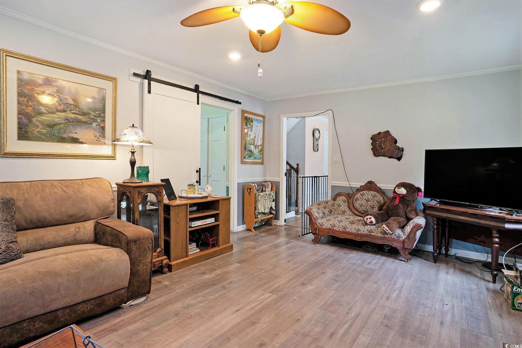 Living room with a barn door, wood finished floors, crown molding, a ceiling fan, and baseboards