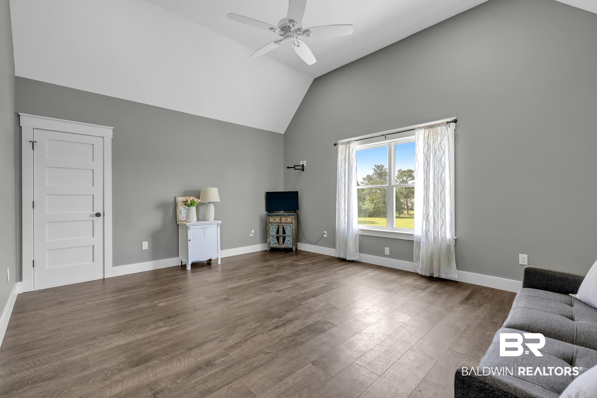 6576 Ranch Loop Road East Grand Bay, AL 36541 - Photo 25 of 39 Vaulted ceiling room featuring wood-finish flooring, a ceiling fan, a prominent window, a white paneled door, and neutral wall tones