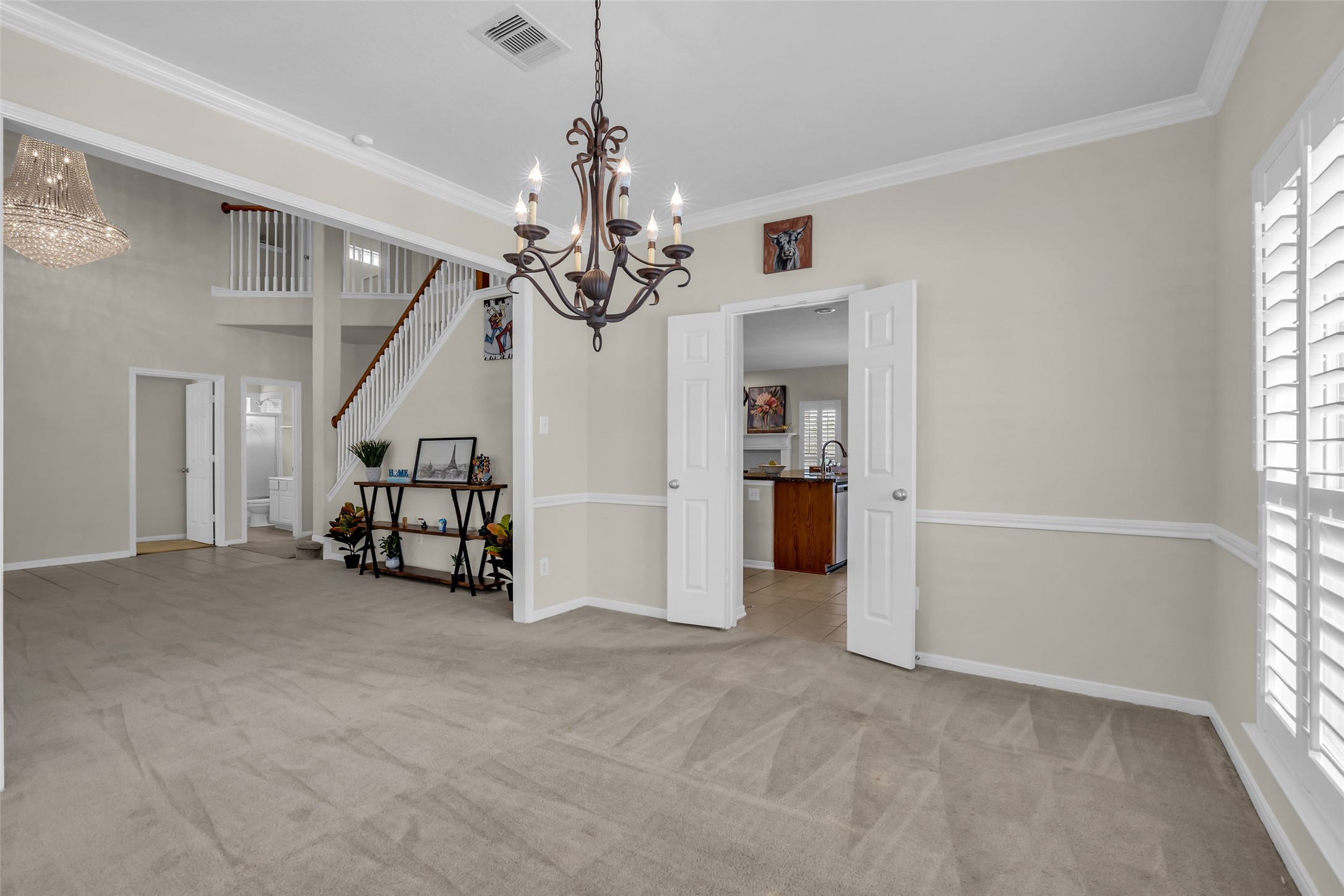 6807 Chapelfield Lane Houston, TX 77049 - Photo 11 of 42 This photo features a spacious, light-filled living area with neutral walls and carpet. It showcases a decorative chandelier, double doors leading to a kitchen, and a staircase with a wooden banister. Large windows with shutters provide ample natural light.