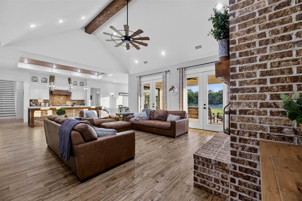 985 Vest Lane Pilot Point, TX 76258 - Photo 17 of 40 Open-concept living space featuring wood-finish flooring, a vaulted ceiling with exposed beam, an exposed brick fireplace, and double glass doors leading to an exterior patio