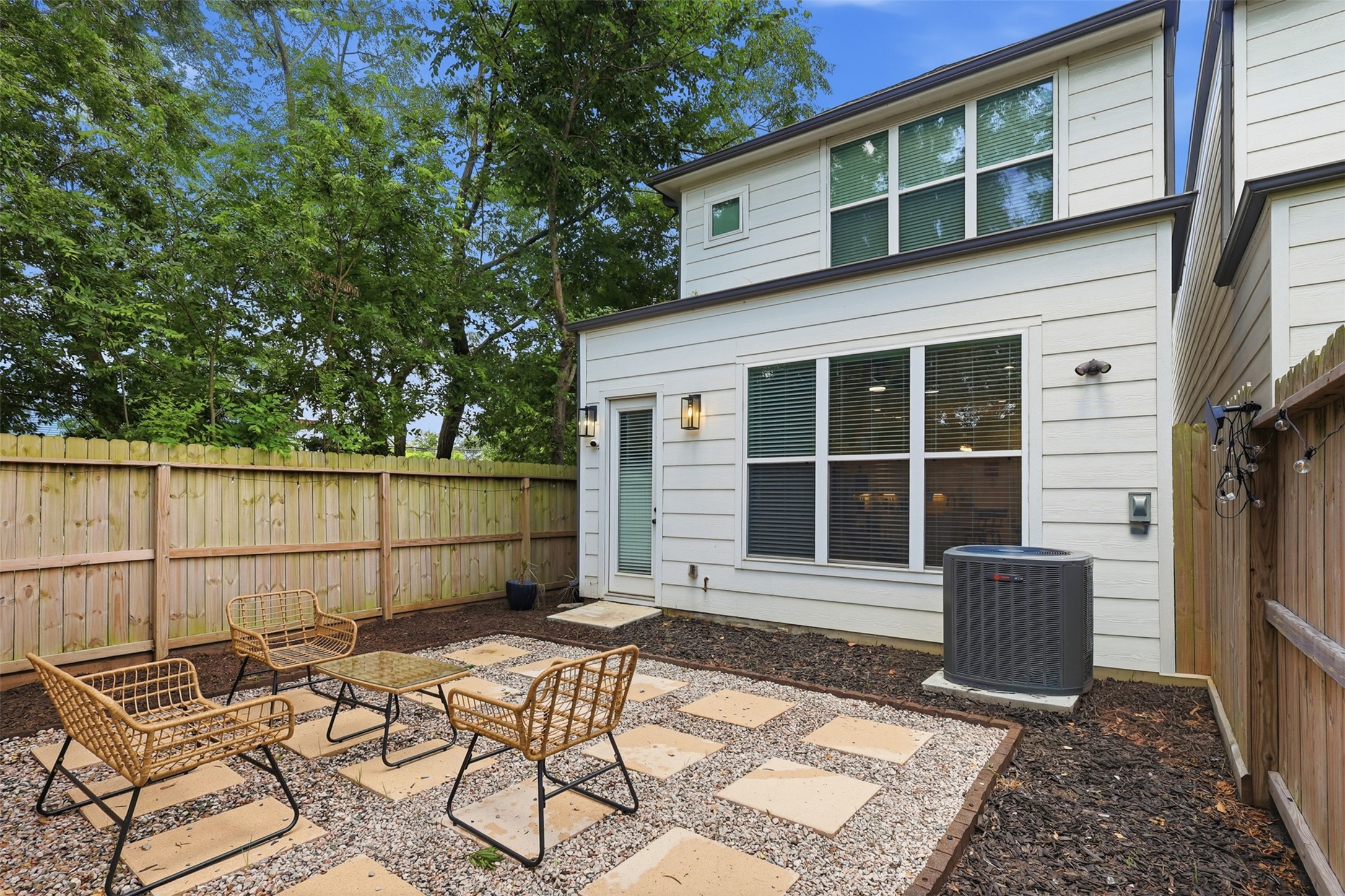 6833 Paris Street Houston, TX 77021 - Photo 23 of 23 This photo showcases a cozy backyard patio area featuring modern outdoor seating on a gravel and paver surface. The space is enclosed by a wooden fence, offering privacy, and is bordered by trees, providing a natural backdrop. The exterior of the home has large windows and a small air conditioning unit is visible.