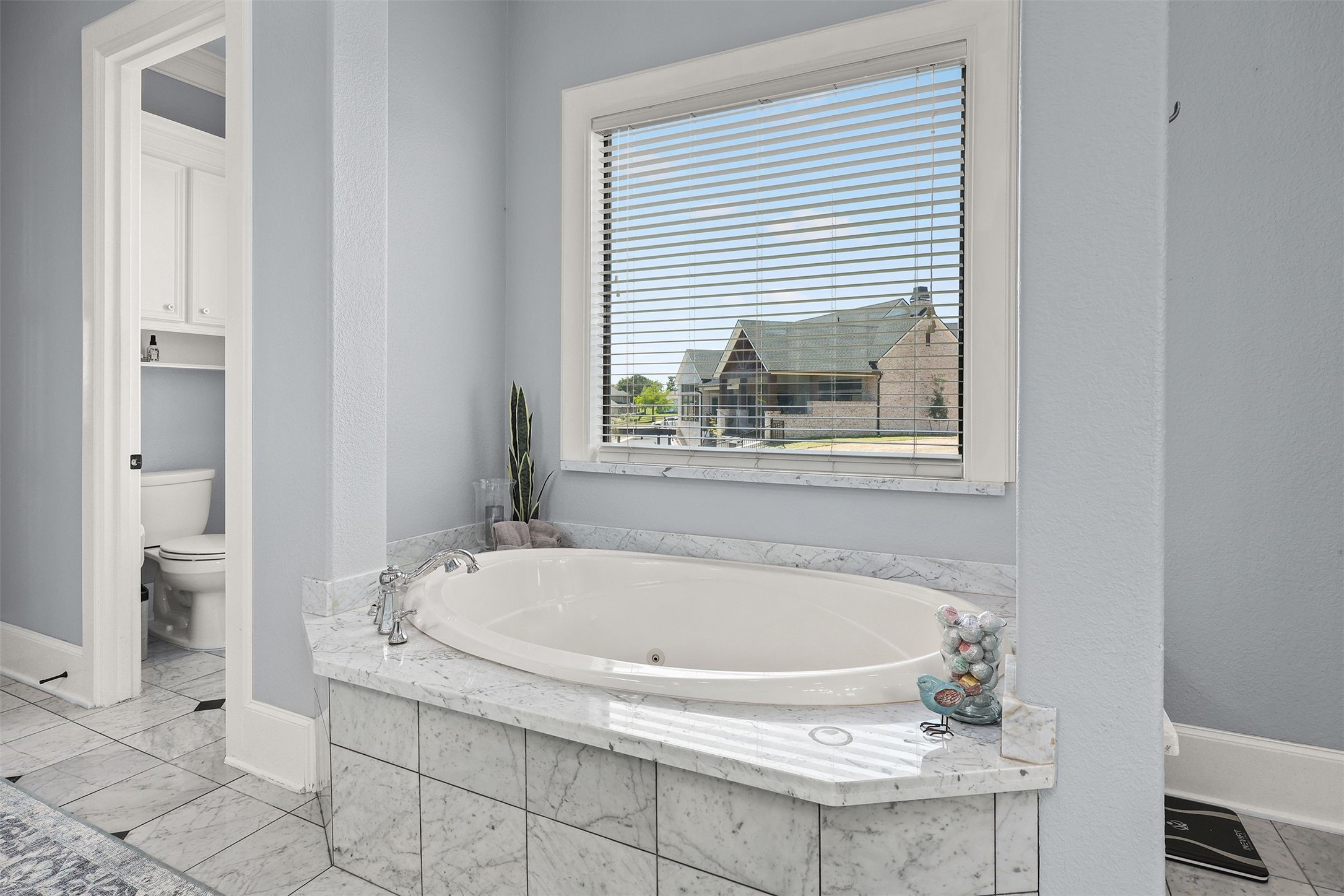 10896 Decatur Street Willis, TX 77318 - Photo 34 of 50 This elegant bathroom features a luxurious marble-tiled soaking tub beneath a window, inviting natural light and views, with a separate toilet area boasting convenient storage cabinets.