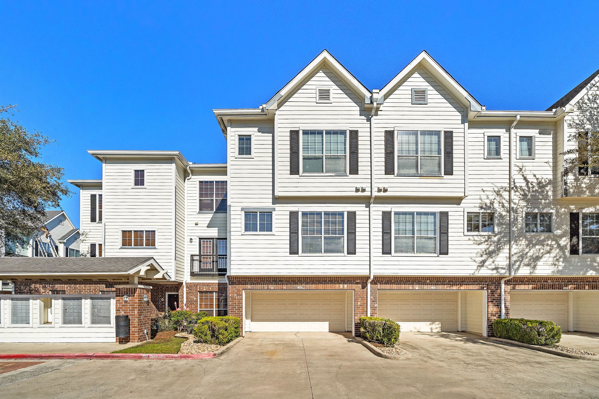 This photo shows a modern townhouse/condominium complex with crisp white siding and brick accents. The building features multiple stories, large windows for natural light, and attached garages. The driveway is spacious, and there are small landscaped areas with shrubs.