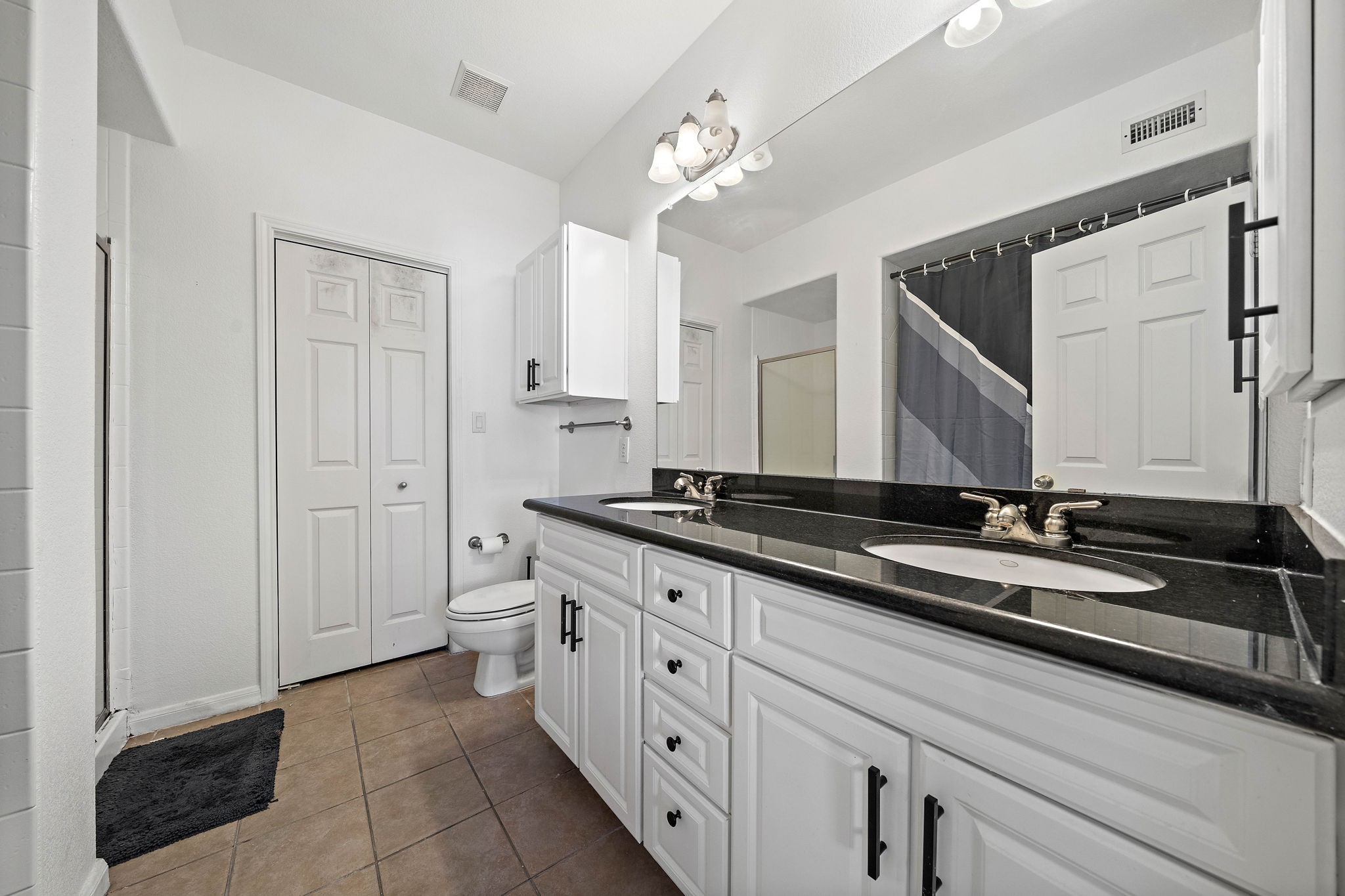 9200 Westheimer Road, Unit 203 Houston, TX 77063 - Photo 6 of 10 This bathroom features a modern design with a double vanity, black countertops, and white cabinetry. It includes a shower with a curtain, tiled flooring, and ample lighting, offering a clean and functional space.