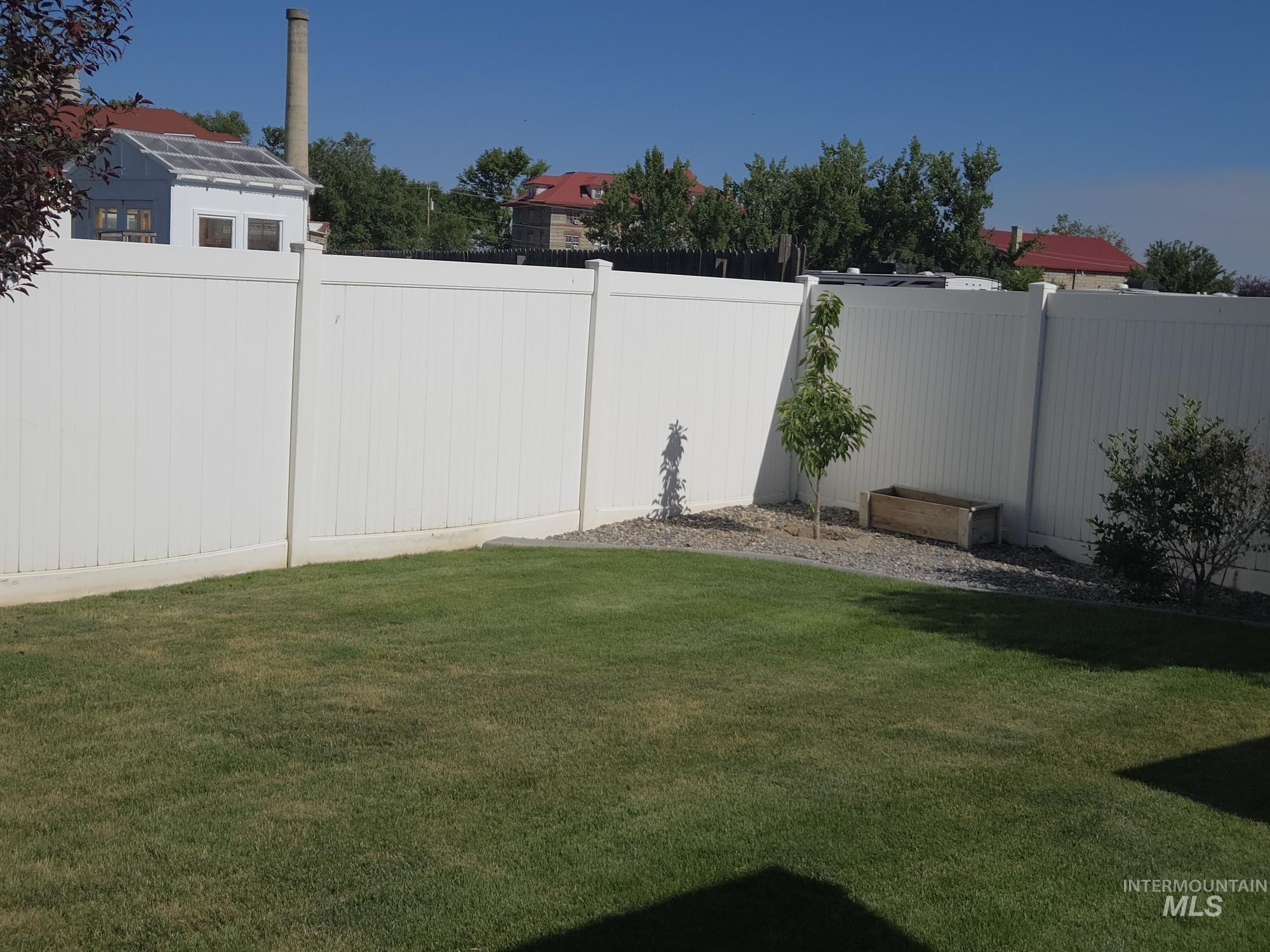 2480 West 9th Street Weiser, ID 83672 - Photo 15 of 17 Expansive lawn area enclosed by a tall white privacy fence, featuring a gravel border with a young tree and shrub