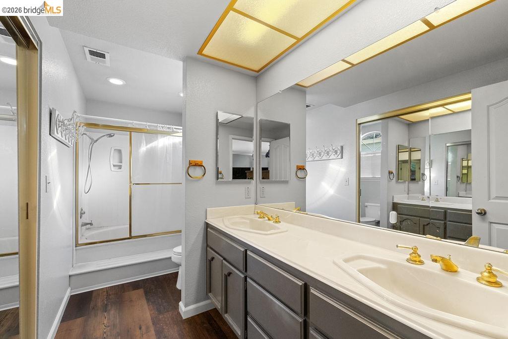 724 Winterside Circle San Ramon, CA 94583 - Photo 22 of 43 Bathroom featuring a dual vanity with gold-tone fixtures, wood-finish flooring, a full-width mirror, and a bathtub with a sliding glass enclosure
