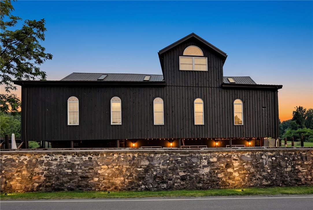 101 Conrad Road Fleetwood, PA 19522 - Photo 1 of 42 A striking modern barn-style home with a metal roof sits atop a rustic stone foundation, illuminated by the twilight glow.