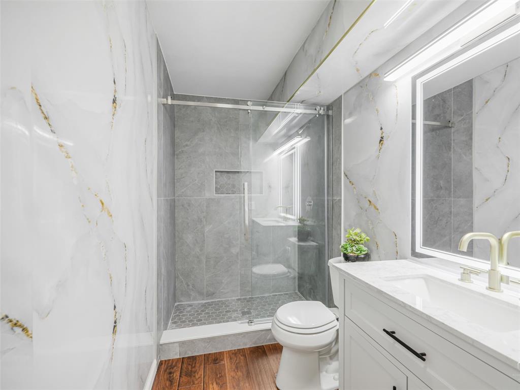 1716 East Parker Road Plano, TX 75074 - Photo 15 of 25 Bathroom featuring white and gold veined wall panels, a glass-enclosed shower with gray tiled walls and a hexagonal tile floor, wood-finish flooring, a white vanity with matte black hardware, and an illuminated mirror