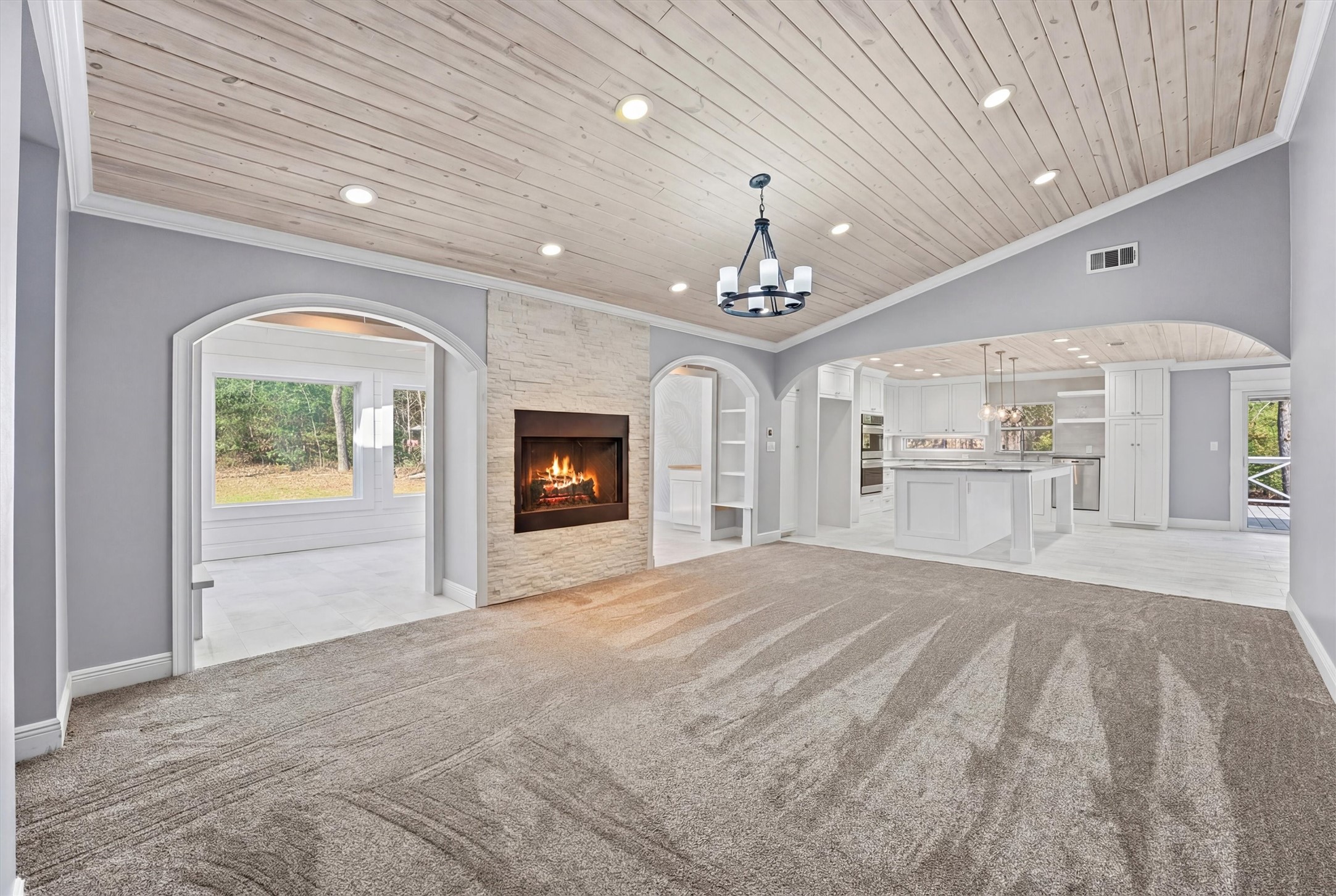 Striking living space showcasing a vaulted wood-plank ceiling, stone fireplace, and elegant archways leading to the custom kitchen and light-filled sunroom—creating a warm and inviting centerpiece of this privately situated home.