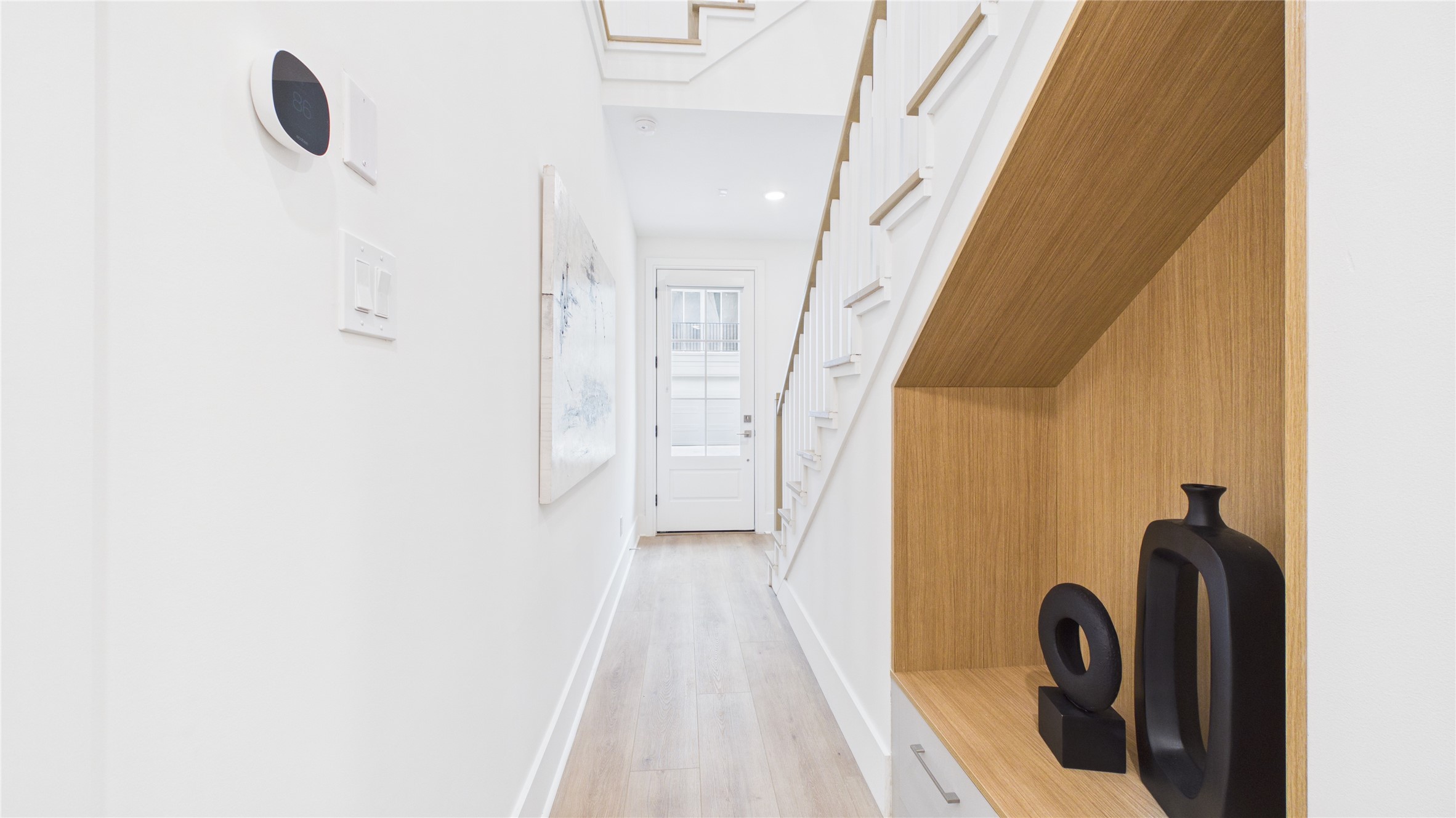 602 Skyland Terrace Way Houston, TX 77009 - Photo 5 of 40 This hallway features a modern, minimalist design with light wood flooring, white walls, and a staircase. There's a built-in wooden shelf with decorative vases, and a door at the end allows natural light to flood in.