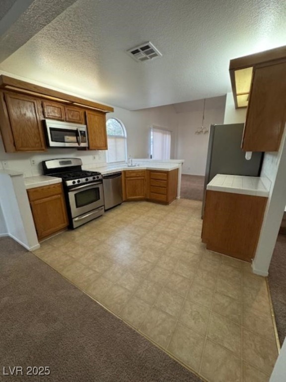 713 Rusty Spur Drive Henderson, NV 89014 - Photo 6 of 15 Kitchen featuring stainless steel appliances, brown cabinets, a textured ceiling, a peninsula, and light carpet