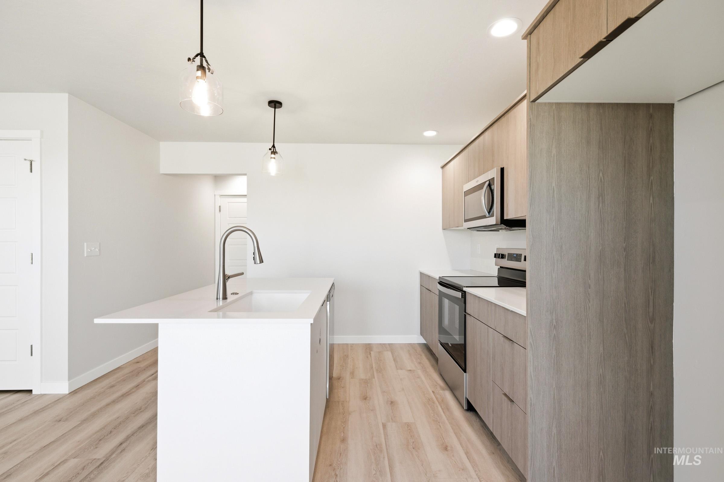 10806 Rutland Street Caldwell, ID 83605 - Photo 7 of 21 Modern kitchen featuring a white island with an integrated sink, wood-finish flooring, light wood-look cabinetry, stainless steel appliances, and pendant lighting
