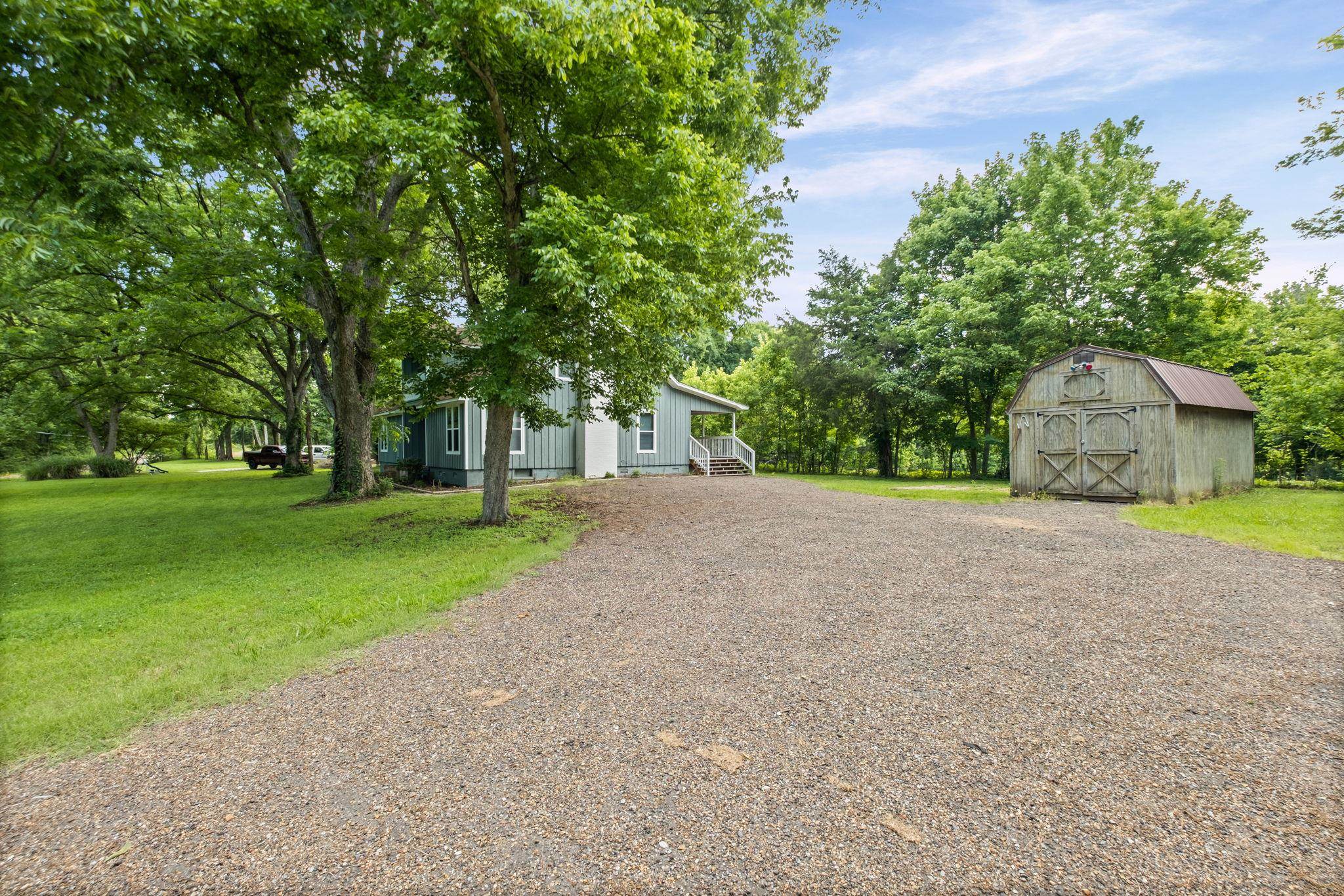 6224 Benjestown Road Memphis, TN 38127 - Photo 5 of 39 Gravel driveway leading to a light blue siding residence with a white porch and steps, surrounded by mature trees, and featuring a rustic barn with a metal roof