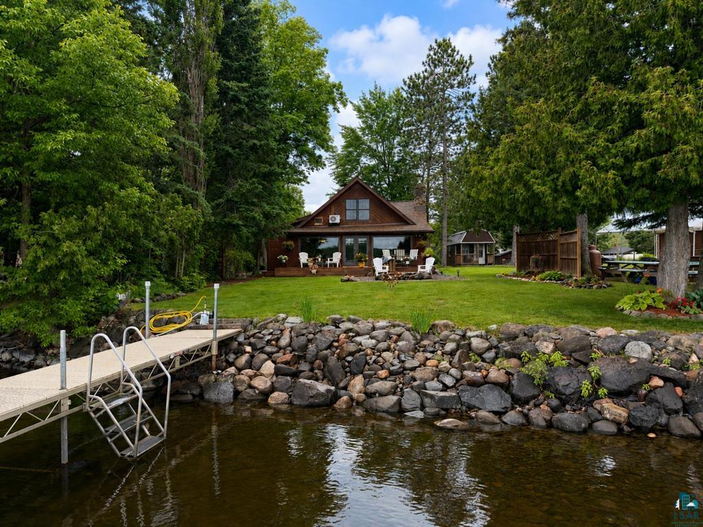 Waterfront property featuring a private dock with ladder, a rock-lined shore, a spacious green lawn, a wood-sided residence with an expansive deck, and mature perimeter trees