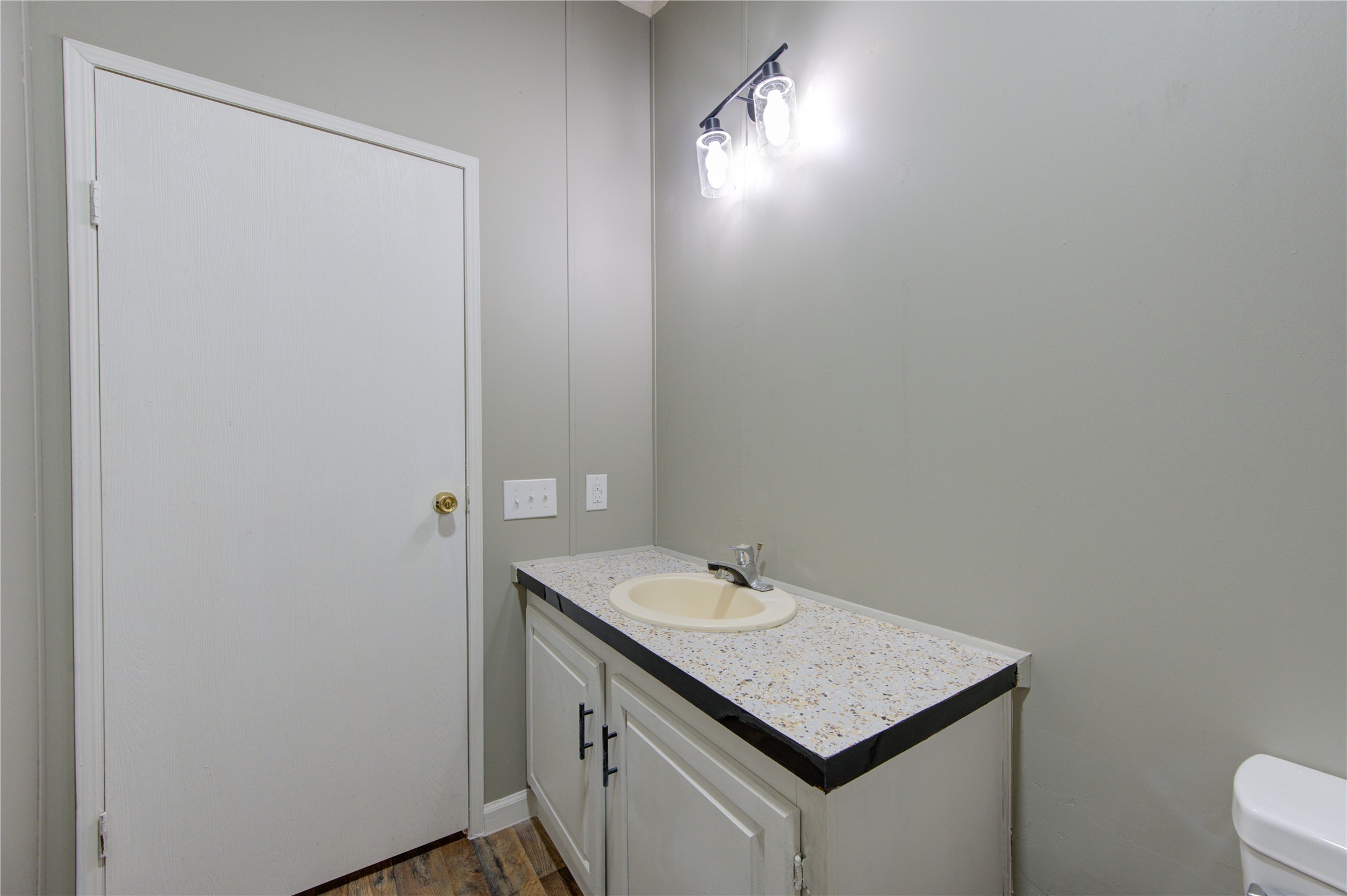 30226 Davis Street Magnolia, TX 77355 - Photo 34 of 50 This is a small bathroom featuring a single sink with a speckled countertop, white cabinetry, and modern lighting. The walls are painted a neutral gray, and the flooring appears to be wood or laminate. There's a white door and visible light switches.