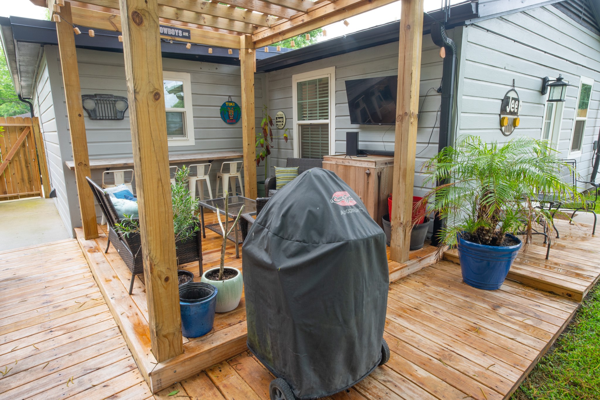 846 South Anderson Street Angleton, TX 77515 - Photo 25 of 30 This photo showcases a cozy outdoor patio area with a wooden deck and pergola. It features comfortable seating, a mounted TV, a covered grill, and potted plants, creating an inviting space for relaxation and entertainment.