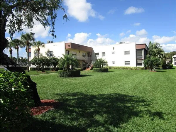 a view of a white house in front of a big yard with plants and large trees