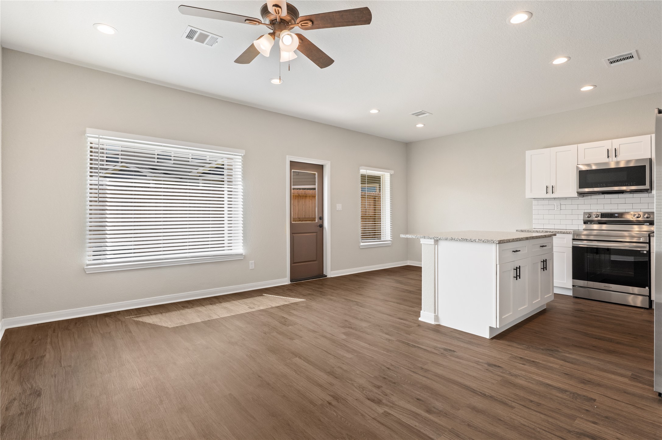 14010 Longstreet Road, Unit 2 Willis, TX 77318 - Photo 24 of 29 This photo showcases a modern, open-concept kitchen and living area with wood flooring, neutral walls, and recessed lighting. The kitchen features stainless steel appliances, white cabinetry, and a central island. A ceiling fan enhances the space, and large windows provide natural light.