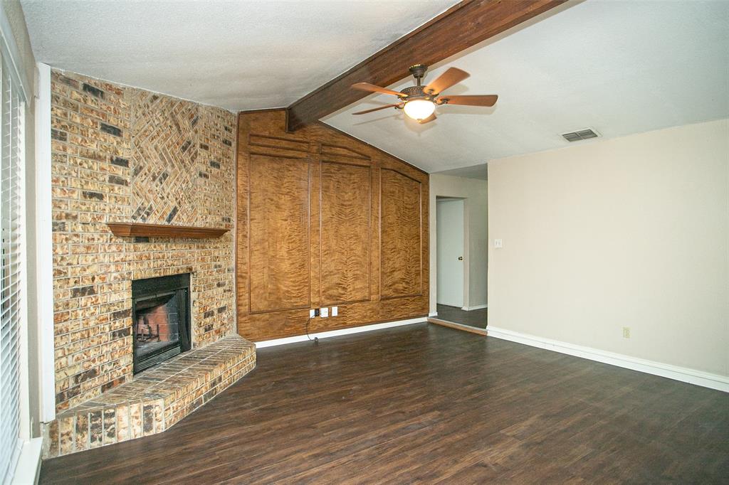 2019 West Nathan Lowe Road Arlington, TX 76017 - Photo 3 of 21 Expansive living area featuring vaulted ceilings with exposed wood beams, a brick fireplace with a hearth and wood mantel, wood-finish flooring, and a pass-through window to the kitchen with a granite-style countertop
