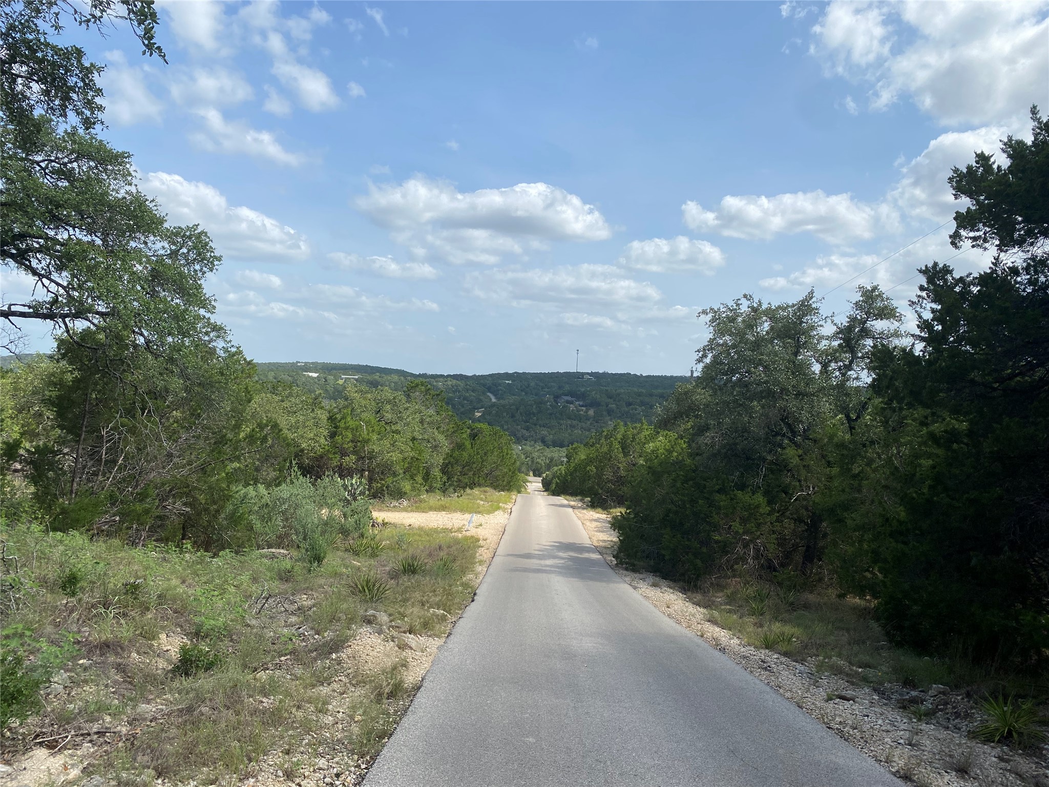 Tbd Whisper Ridge Wimberley, TX 78676 - Photo 2 of 7 A scenic road winds through a verdant landscape, revealing a distant tree-filled valley under a cloud-strewn sky.