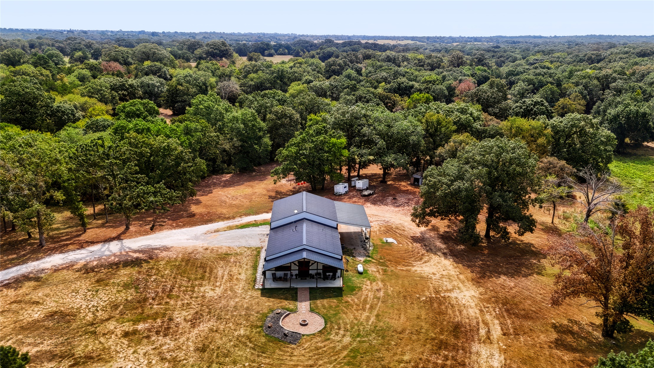 3607 South Half Bank Road Atoka, OK 74525 - Photo 60 of 82 Metal roof property with extensive surrounding tree coverage, a gravel driveway, and a circular paved patio with a fire pit