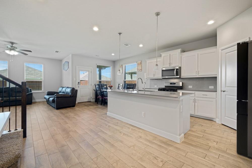 1112 Croxley Way Justin, TX 76247 - Photo 3 of 18 Open-concept living space featuring wood-finish flooring, a kitchen island with pendant lighting, white cabinetry, stainless steel appliances, and multiple windows with blinds