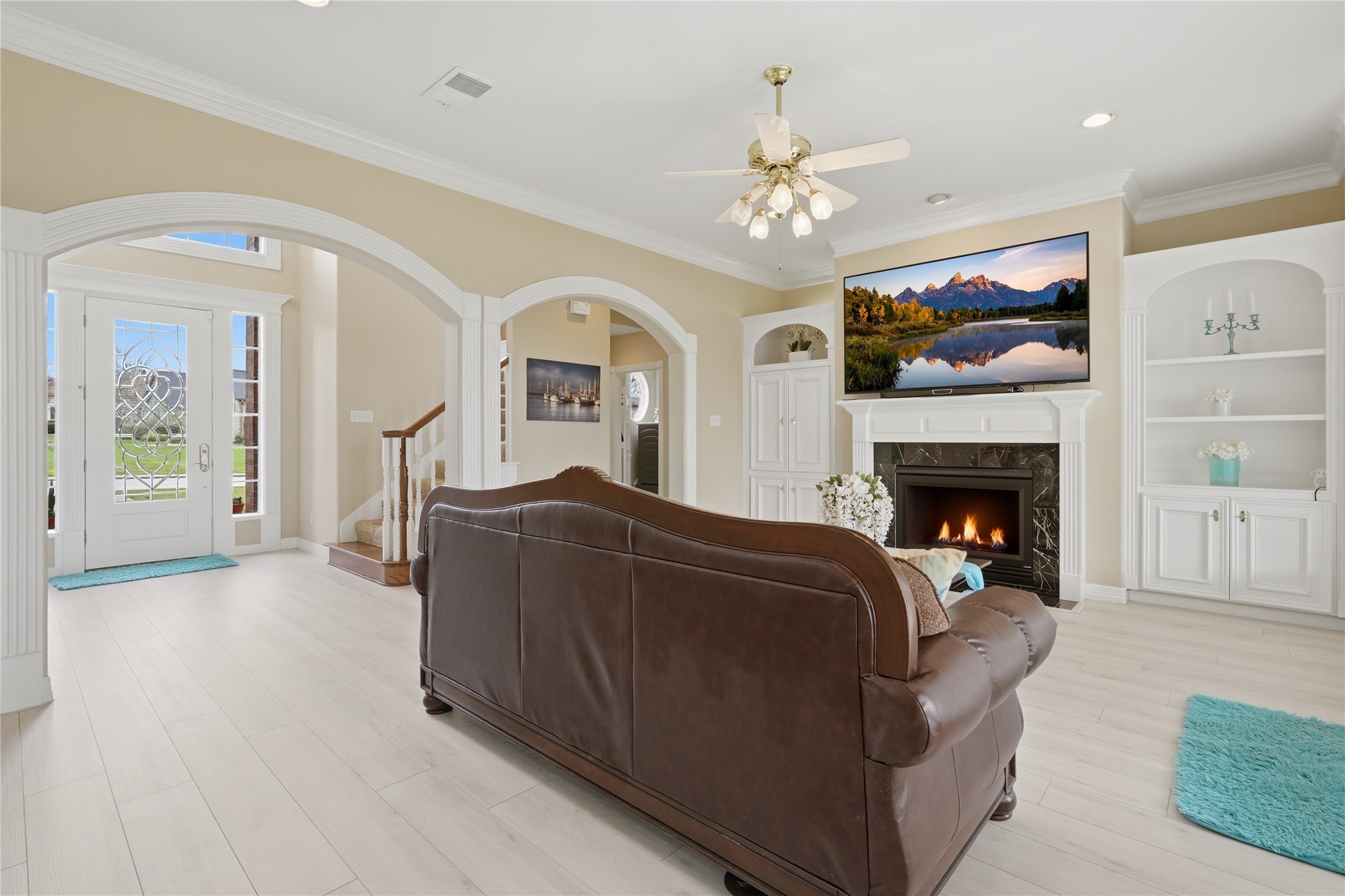 55 Monterrey Road East Montgomery, TX 77356 - Photo 11 of 50 This inviting living room features a cozy fireplace, elegant built-in shelves. It has an open layout with light wood floors, a ceiling fan, and natural light from the adjacent entryway, creating a warm and welcoming atmosphere.
