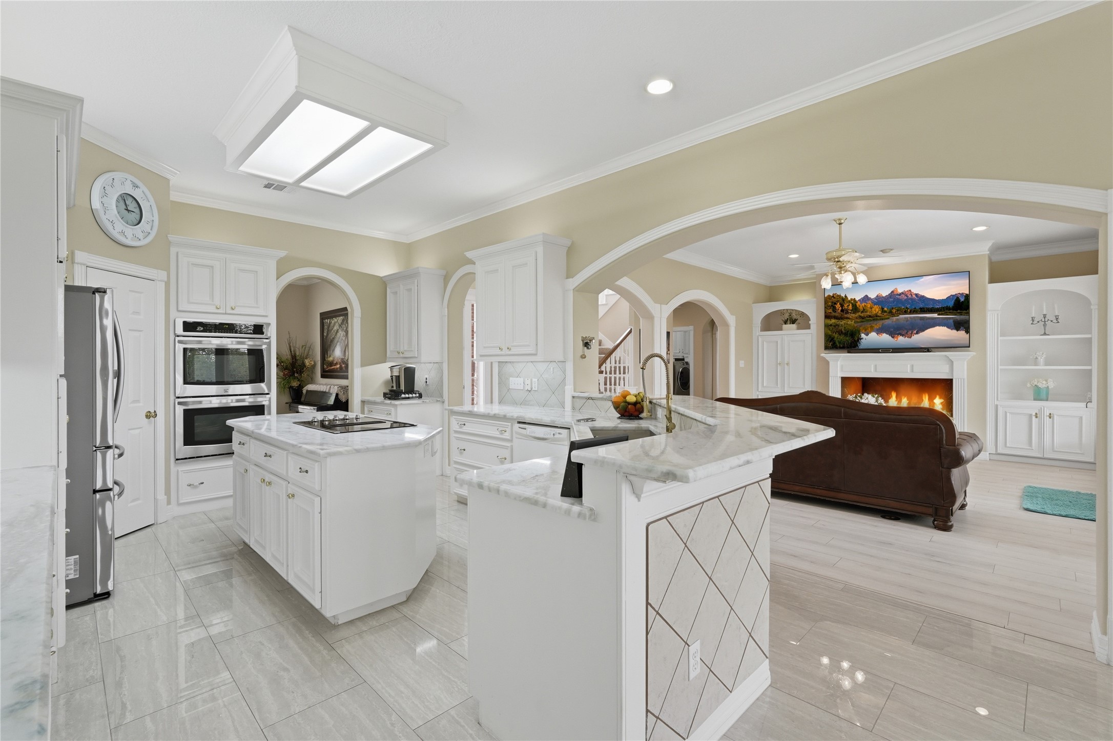55 Monterrey Road East Montgomery, TX 77356 - Photo 16 of 50 This photo showcases a bright, open-concept kitchen and living area. The kitchen features white cabinetry, marble countertops, and modern appliances. It seamlessly flows into a cozy living room with a fireplace, built-in shelves. Arched doorways and tile flooring add elegance to the space.
