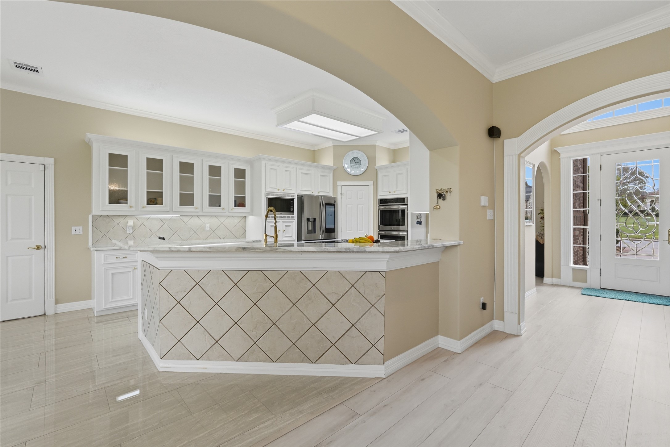 55 Monterrey Road East Montgomery, TX 77356 - Photo 19 of 50 This photo features a bright, spacious kitchen with white cabinetry, stainless steel appliances, and a tiled backsplash. It opens to an inviting entryway with arched doorways and natural light, creating an elegant and welcoming atmosphere.