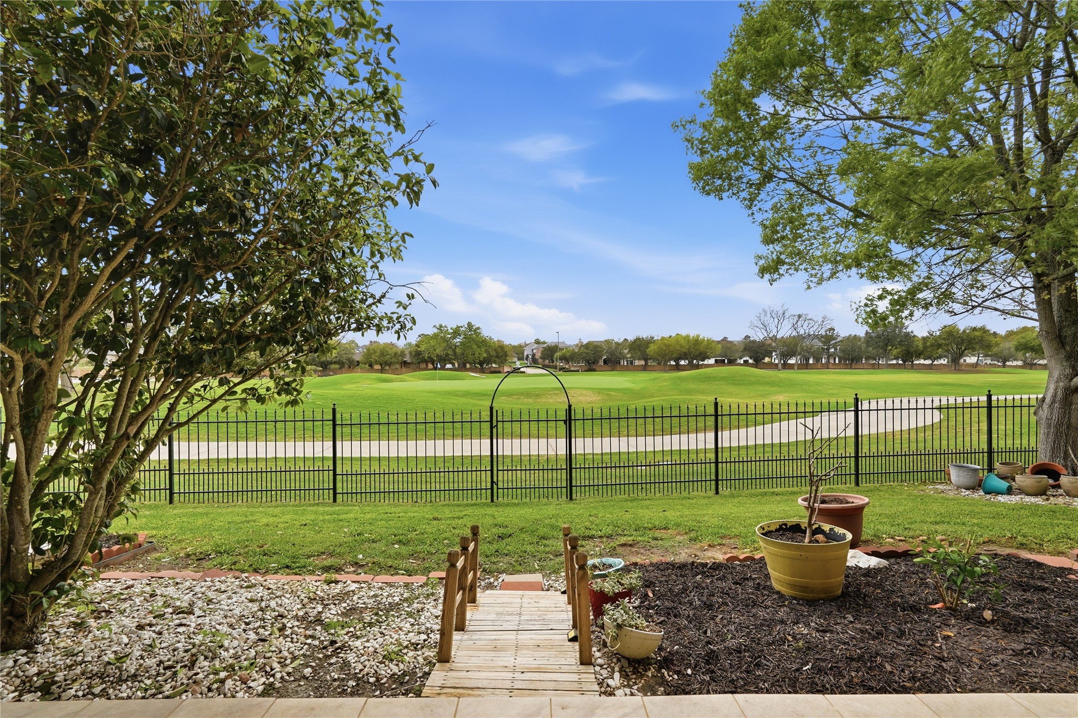 55 Monterrey Road East Montgomery, TX 77356 - Photo 43 of 50 This photo showcases a backyard with a beautifully landscaped garden and a pathway leading to a wrought iron fence. Beyond the fence is a well-maintained golf course, offering expansive green views under a clear blue sky.