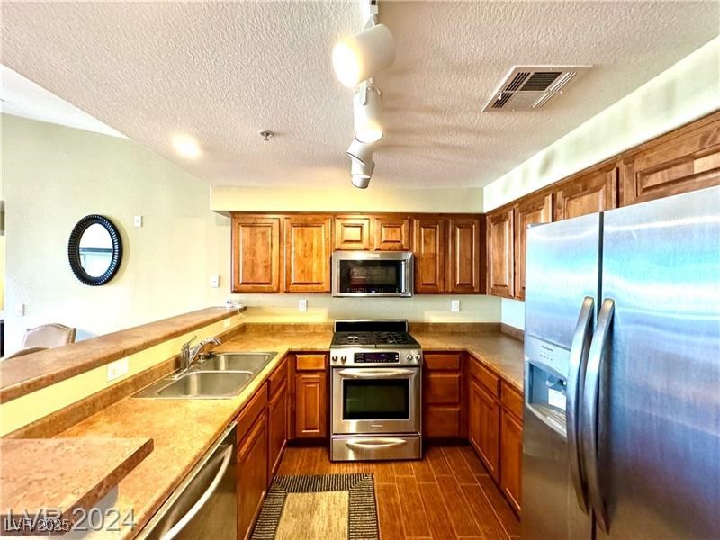 Kitchen featuring appliances with stainless steel finishes, brown cabinetry, dark wood-style flooring, a textured ceiling, and a peninsula