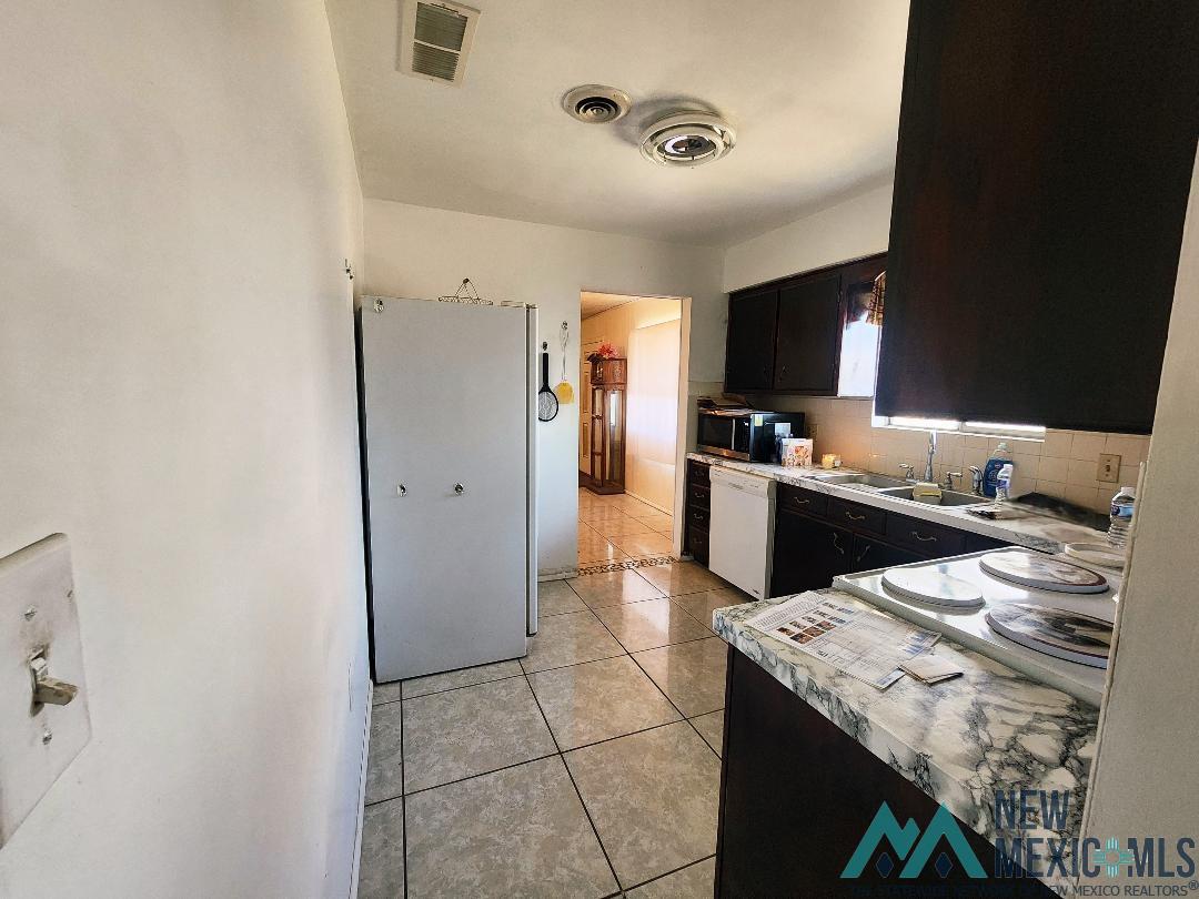 Kitchen featuring light countertops, light tile patterned floors, dark cabinetry, white appliances, and decorative backsplash