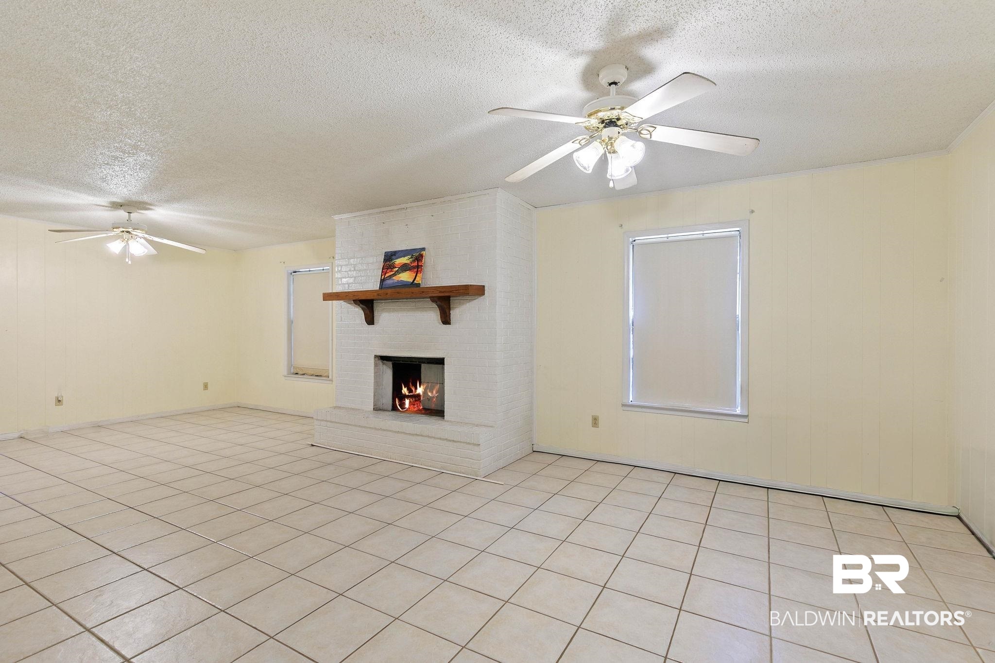 13653 Roberts Road Loxley, AL 36551 - Photo 10 of 33 Spacious room featuring a white brick fireplace with a wood mantel, light-toned tile flooring, and two ceiling fans with integrated lighting