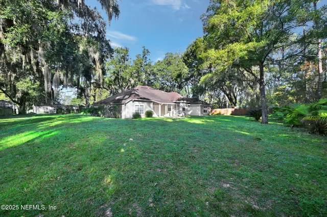 a view of a white house in front of a big yard with plants and large trees