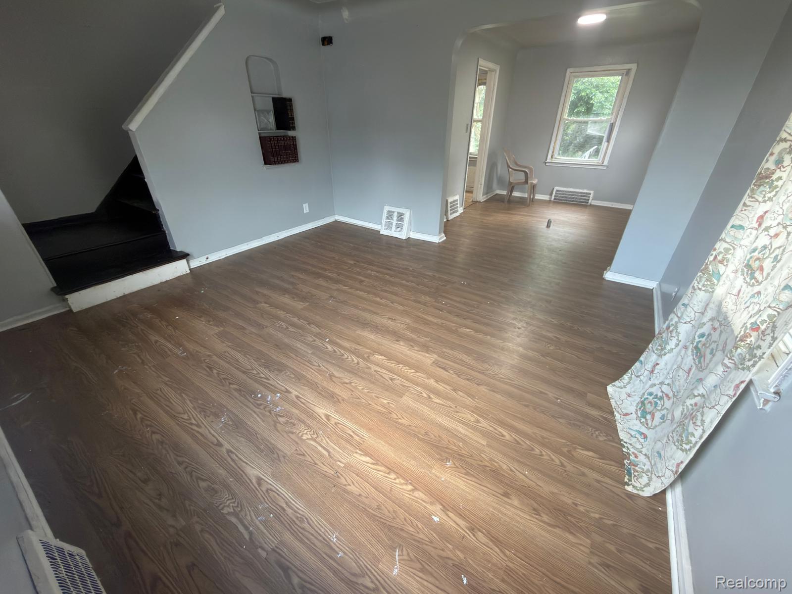 8837 Evergreen Road Detroit, MI 48228 - Photo 8 of 16 Spacious room featuring wood-finish flooring, light gray walls, white baseboards, a visible staircase, and a window providing natural illumination